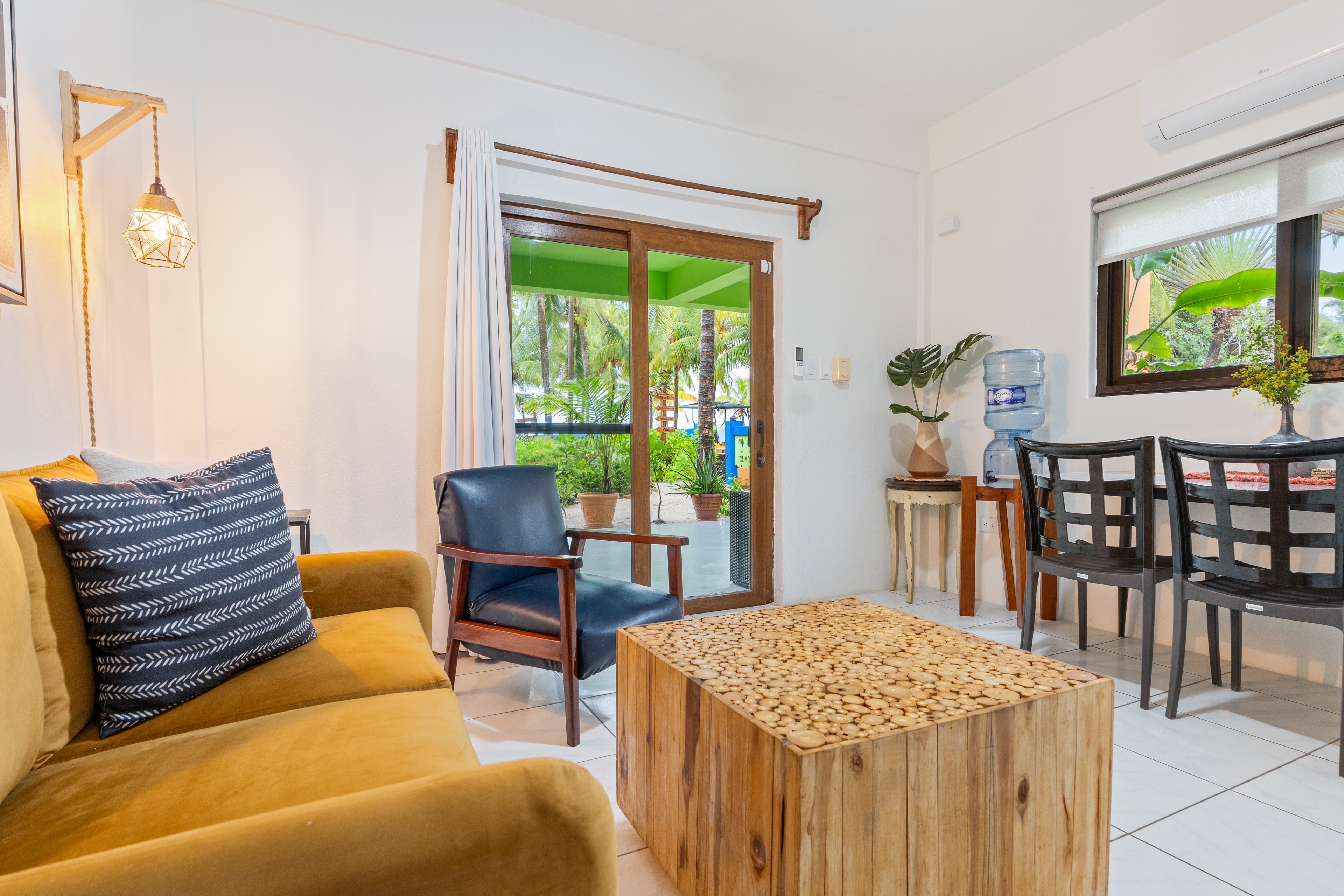 A bright living area featuring an ochre velvet sofa with accent pillows, a solid wood cube coffee table, and a dark leather armchair. The room includes a dining table with black chairs, a water cooler, and a sliding glass door that provides a view of a lush green outdoor area.