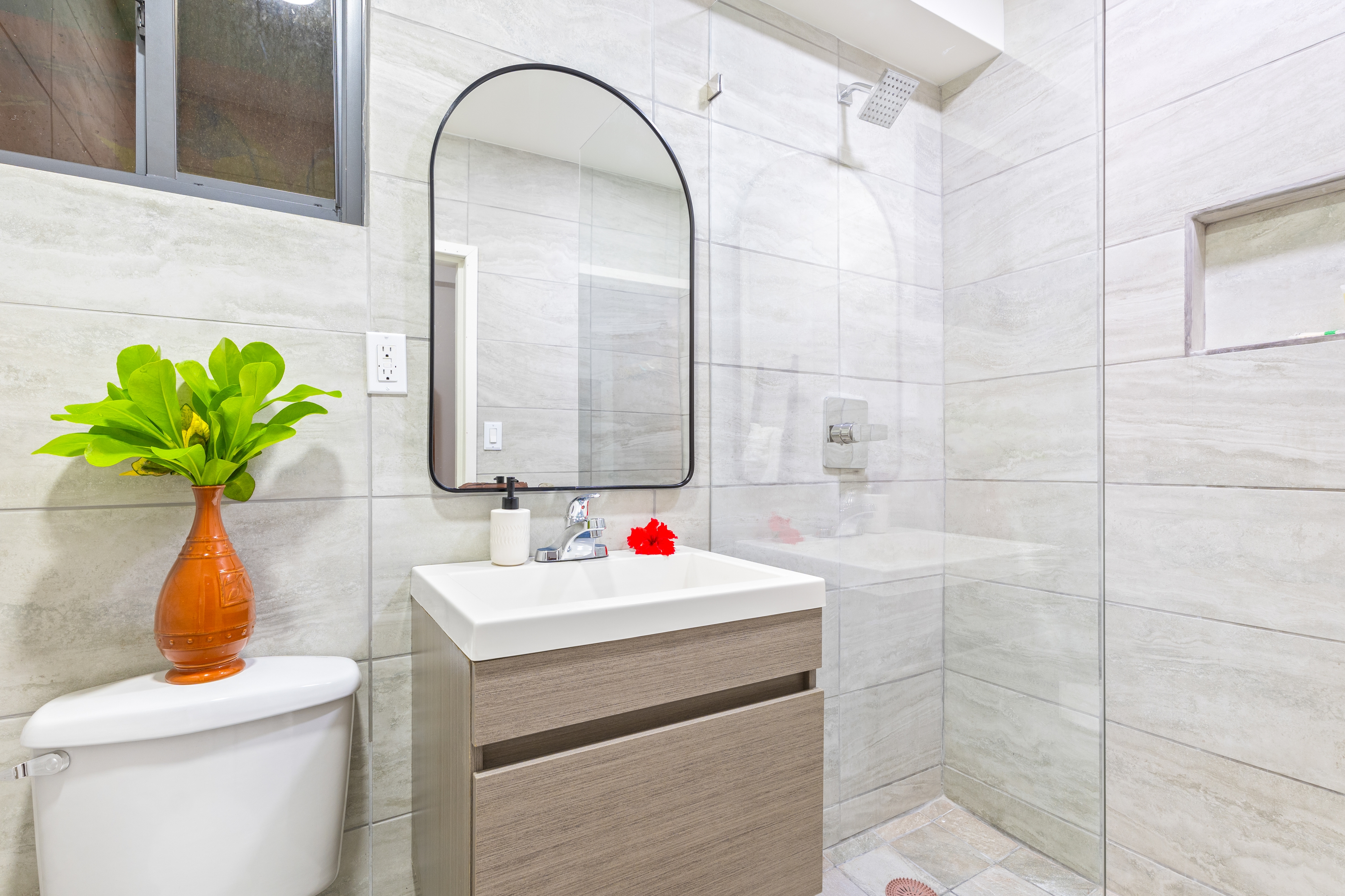 A modern bathroom featuring light gray tiled walls and a glass-enclosed walk-in shower. A floating wood-grain vanity with a white vessel sink is centered under an arched, black-framed mirror, while a white toilet sits to the left, decorated with a vibrant orange vase holding green foliage.