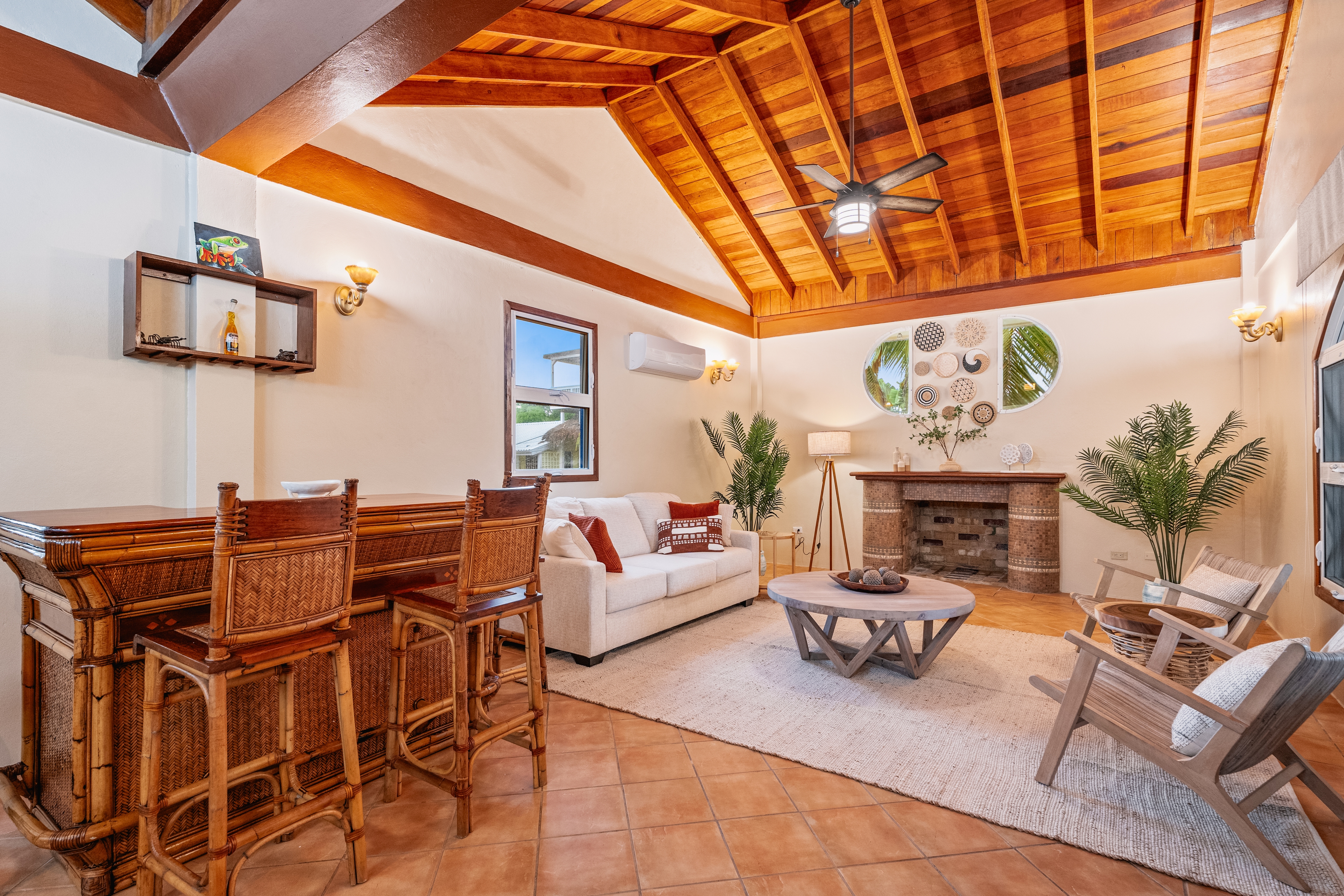 A spacious living room featuring a high, vaulted wooden ceiling with a central fan and terracotta tile flooring. The space is furnished with a bamboo-style bar and matching high-backed stools, a white sofa with red accent pillows, and two wooden lounge chairs centered around a round coffee table. A decorative fireplace mantle sits against the far wall, flanked by indoor plants and topped with circular wall art.