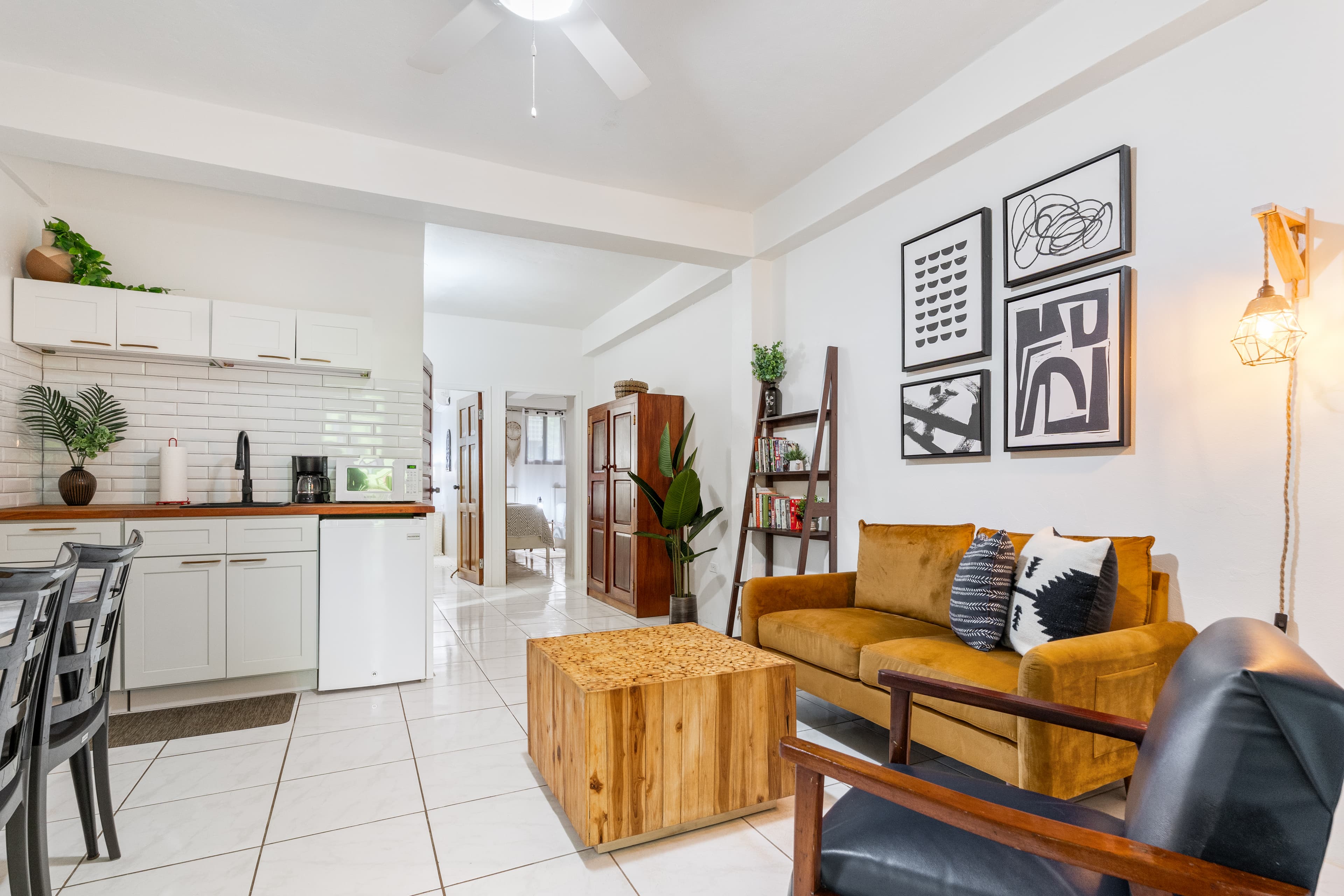 A modern open-concept living area and kitchenette featuring white-tiled floors and bright white walls. The living space includes an ochre velvet sofa, a solid wood cube coffee table, a dark leather armchair, and a decorative wooden ladder shelf. To the left, a small kitchenette displays white cabinetry, a wood-topped counter with a coffee maker and mini-fridge, and four pieces of black-and-white abstract art hanging on the wall.