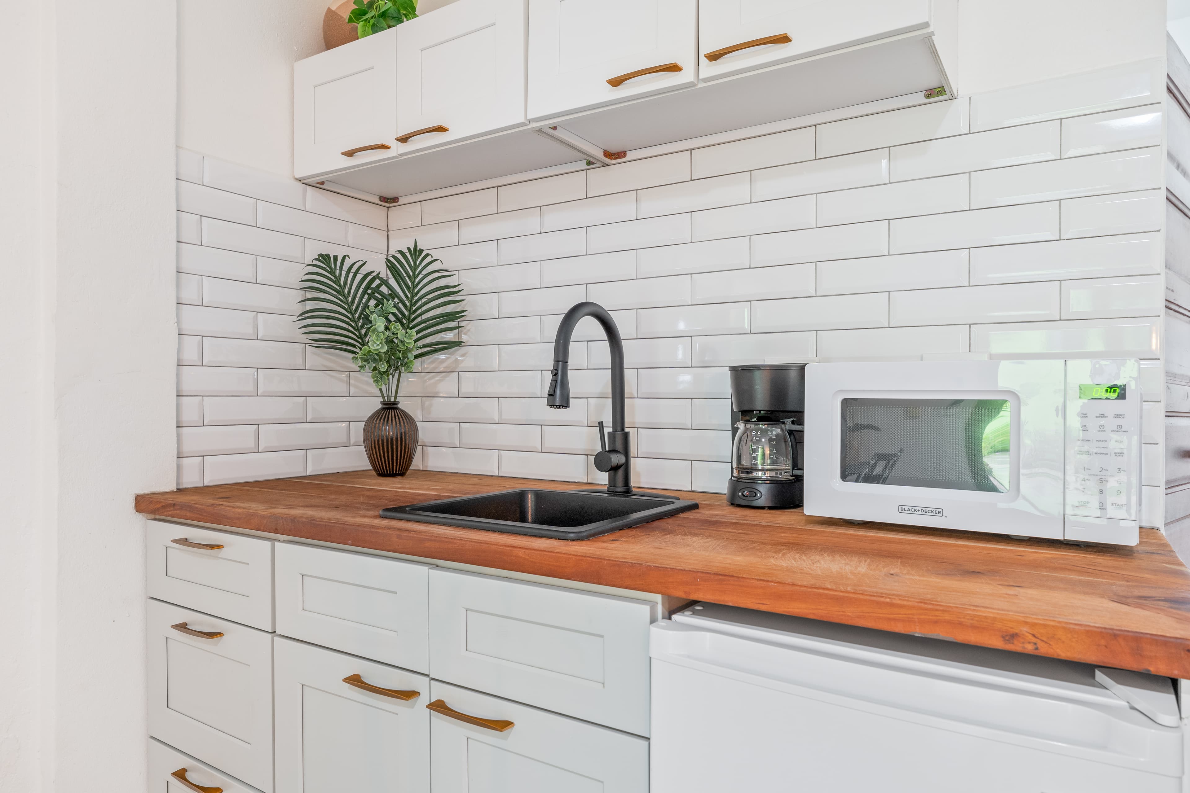 A close-up view of a small kitchenette featuring white shaker-style cabinetry with brown handles and a dark wood countertop. The space is equipped with a white microwave, a black drip coffee maker, a black sink with a matching gooseneck faucet, and a small white refrigerator. A white subway tile backsplash serves as the background, decorated with a small brown vase holding green palm fronds.