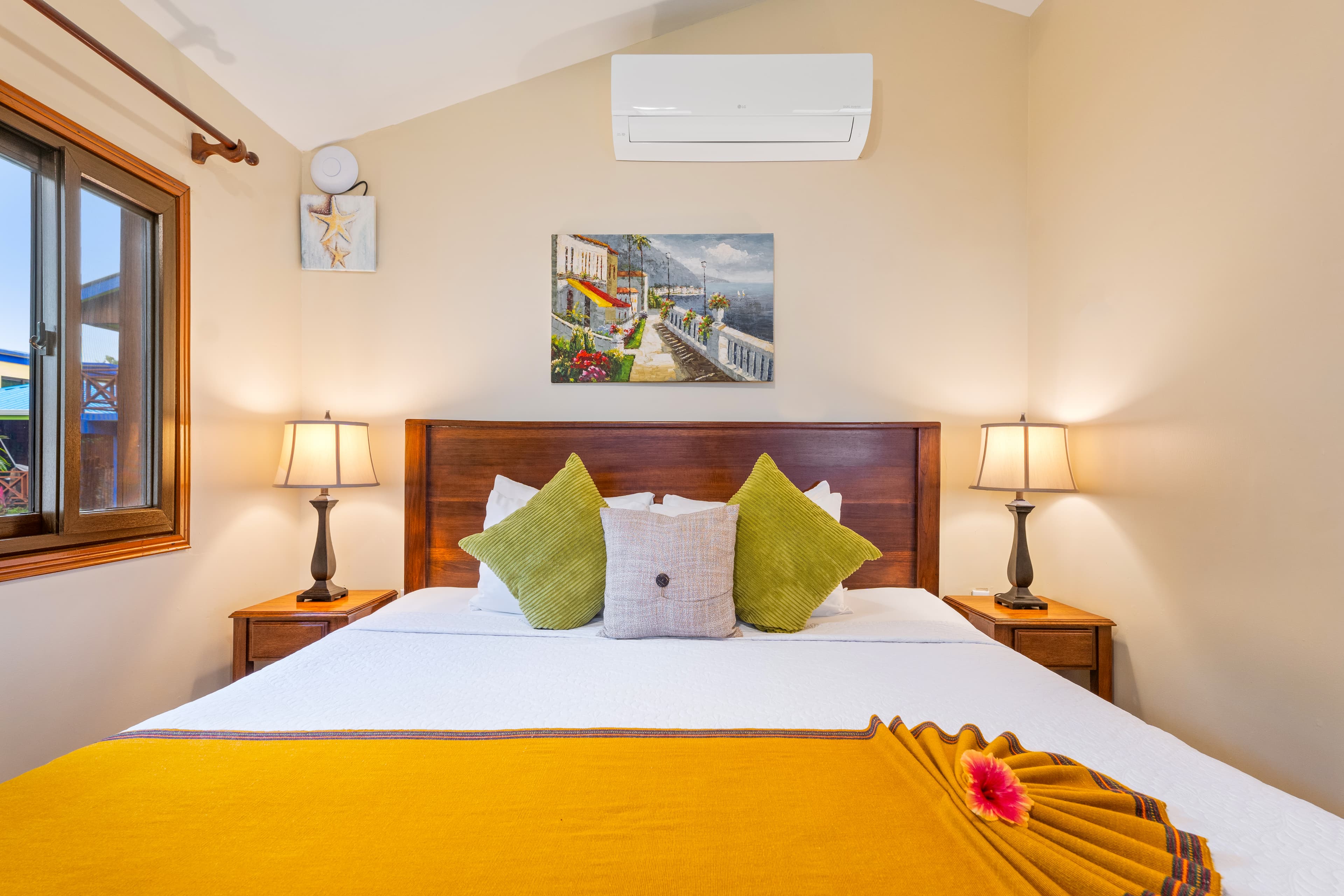 A bedroom with light beige walls and a vaulted ceiling featuring a king-size bed with white linens and a vibrant mustard-yellow runner. The bed is styled with two lime-green pillows and a center grey patterned pillow, flanked by two wooden nightstands with classic lamps. A colorful coastal painting hangs on the wall above the headboard, and a window on the left looks out toward tropical greenery.