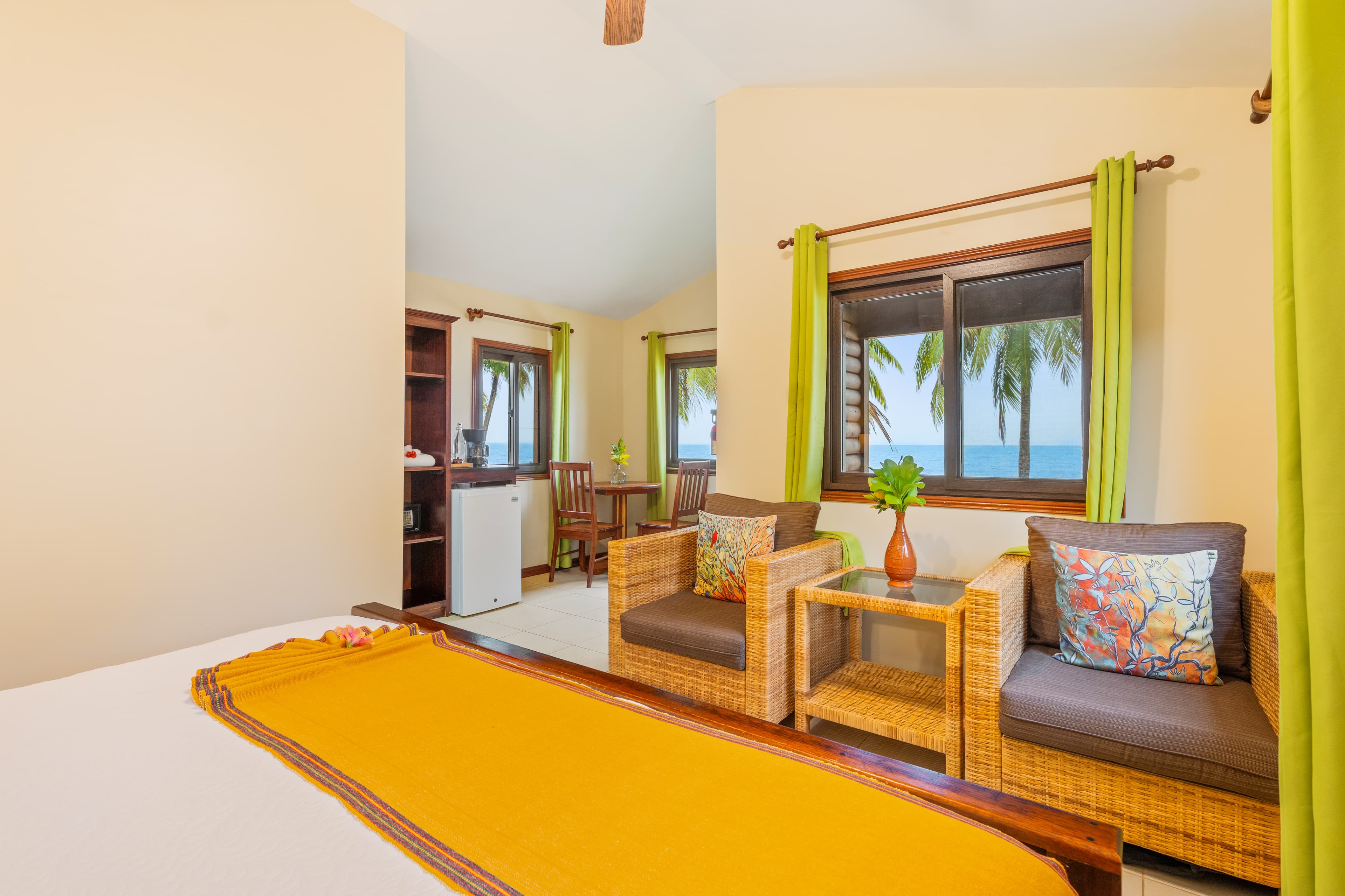 A cozy bedroom featuring a vaulted ceiling and beige walls, with a focus on two wicker armchairs paired with colorful patterned cushions and a small wooden side table. In the background, a king-size bed is styled with a vibrant mustard-yellow runner, while a window with lime-green curtains offers a view of the tropical coast.