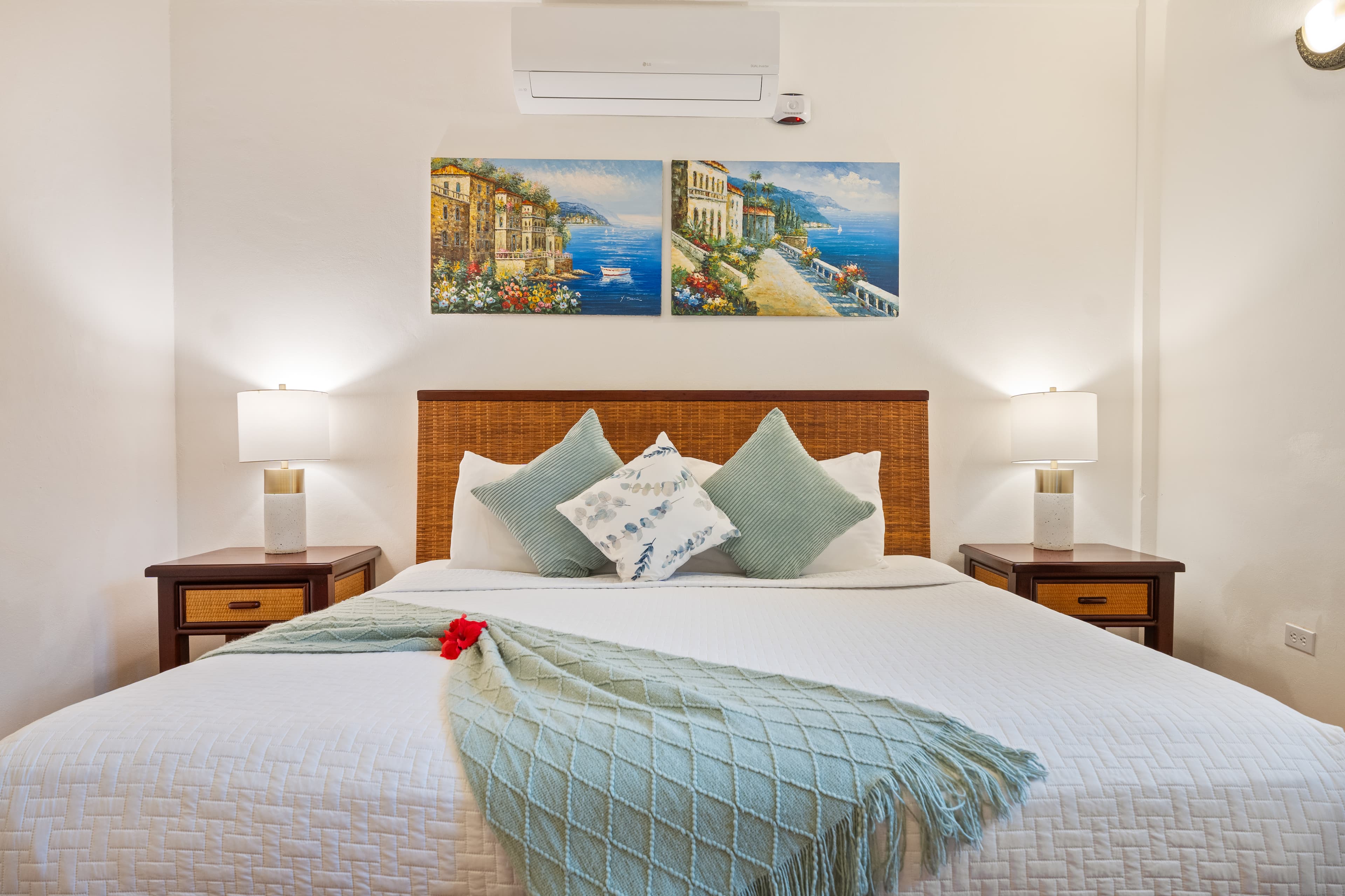 A well-lit bedroom featuring a king-size bed with white linens, seafoam green accent pillows, and a matching throw. Two wooden nightstands with white lamps flank the bed, and two framed coastal paintings hang above the wooden headboard.