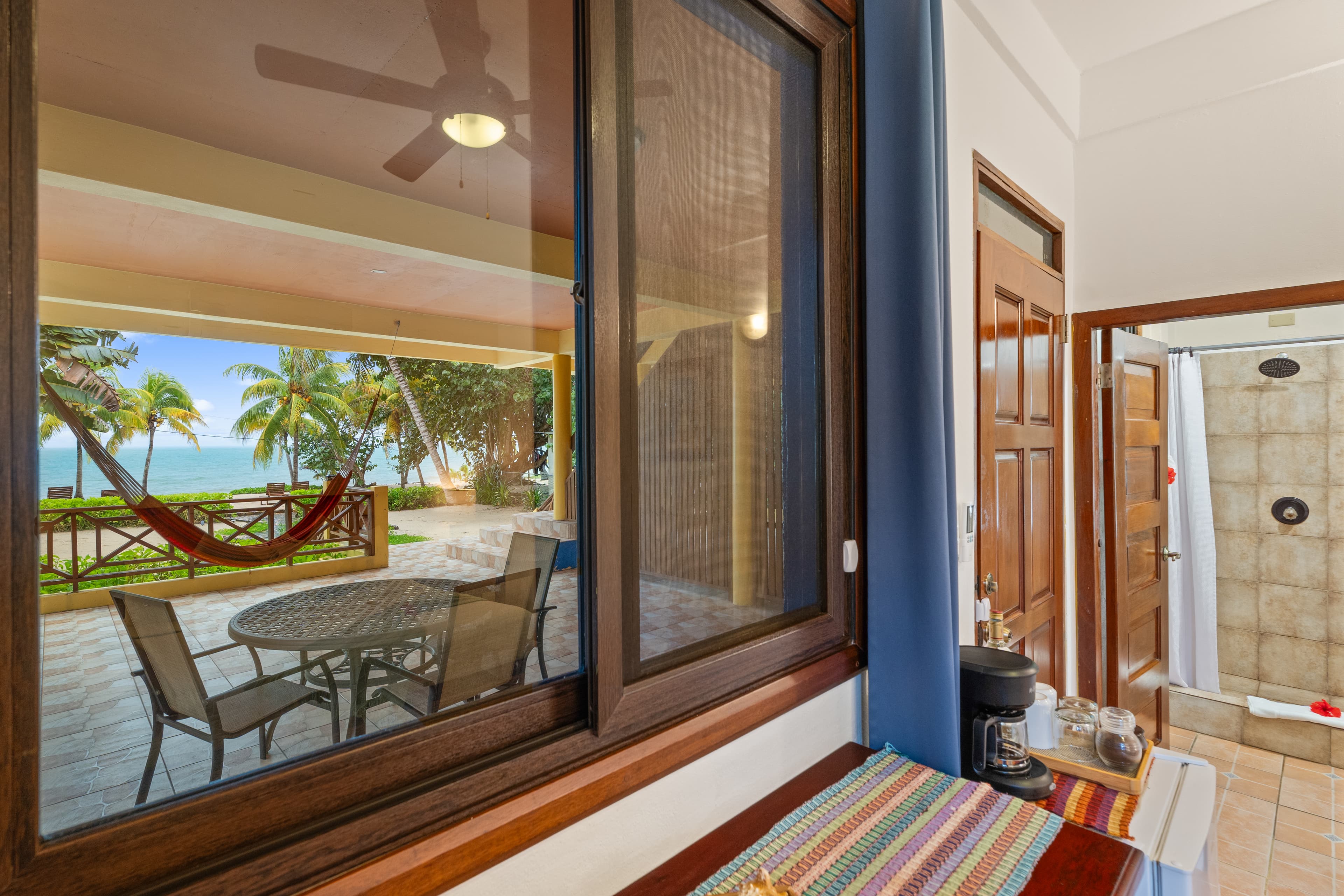 A view through a large window overlooking a private patio with a dining set and a hammock. Beyond the patio, lush tropical palm trees meet the turquoise ocean. Inside, a glimpse of the room shows a coffee maker and an open door leading to a tiled bathroom.