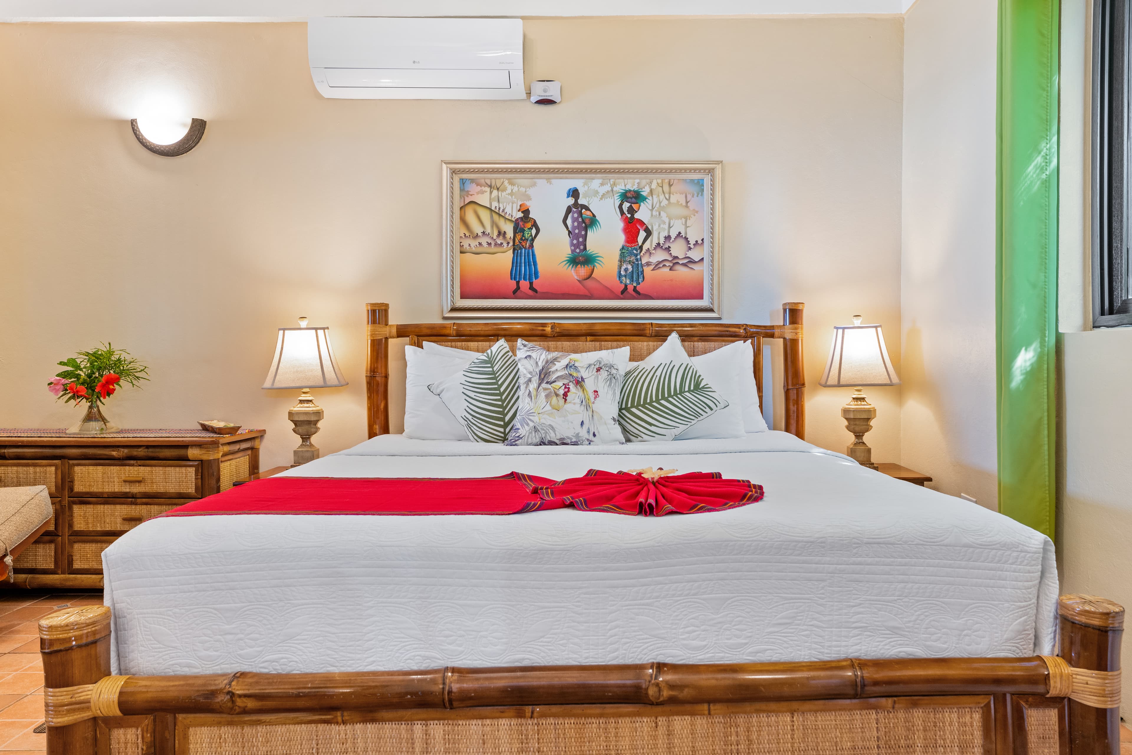 A bedroom with a bamboo-frame bed featuring white linens and a bright red decorative runner. Above the headboard hangs a colorful painting of three women in traditional dress, flanked by two nightstands with lamps and a bamboo dresser on the left.