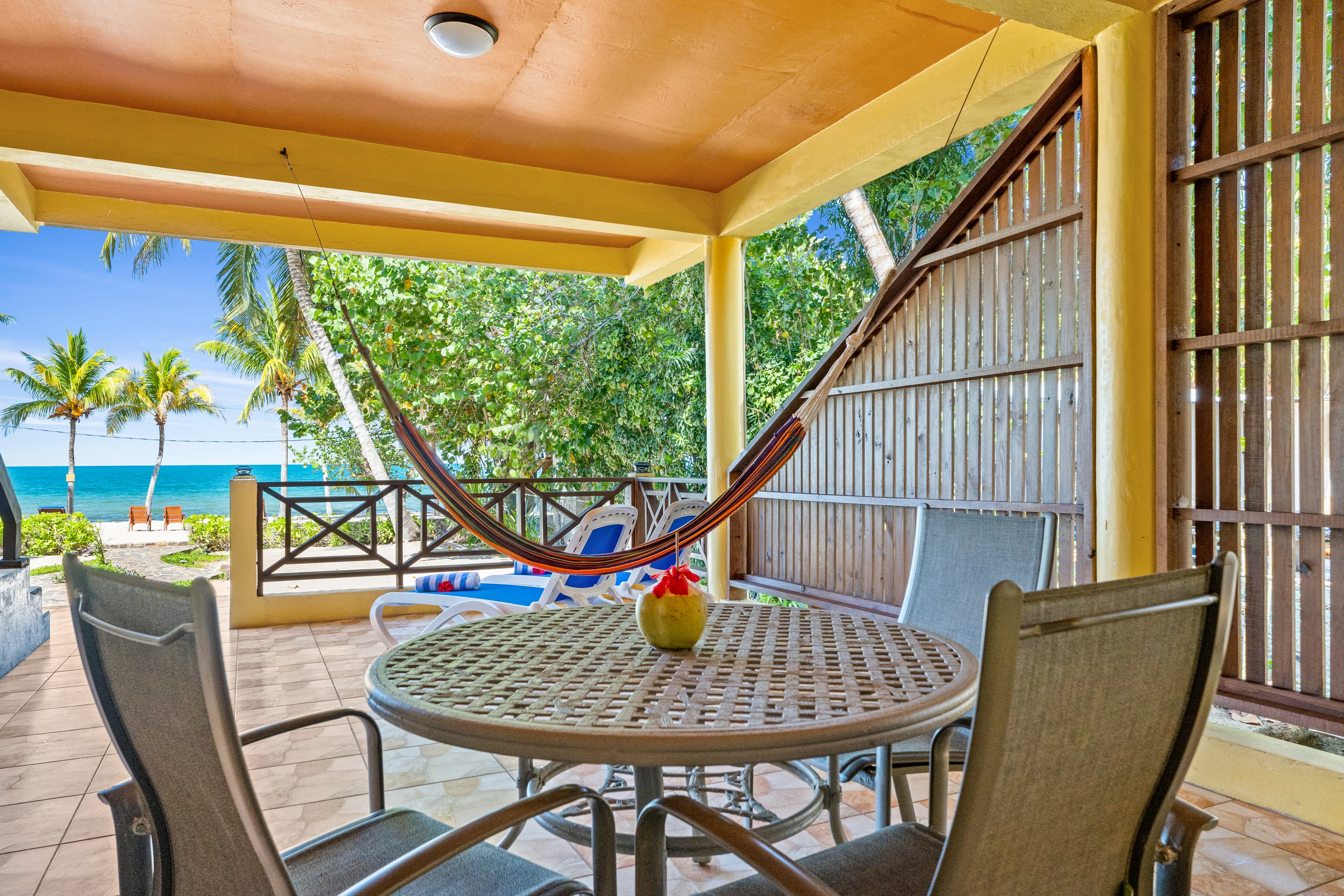 A shaded outdoor patio with a tiled floor features a round, woven-top table surrounded by four mesh-seated chairs. In the background, a multi-colored hammock hangs near a wooden privacy screen, with a view of a lush garden and the bright blue ocean beyond the railing.