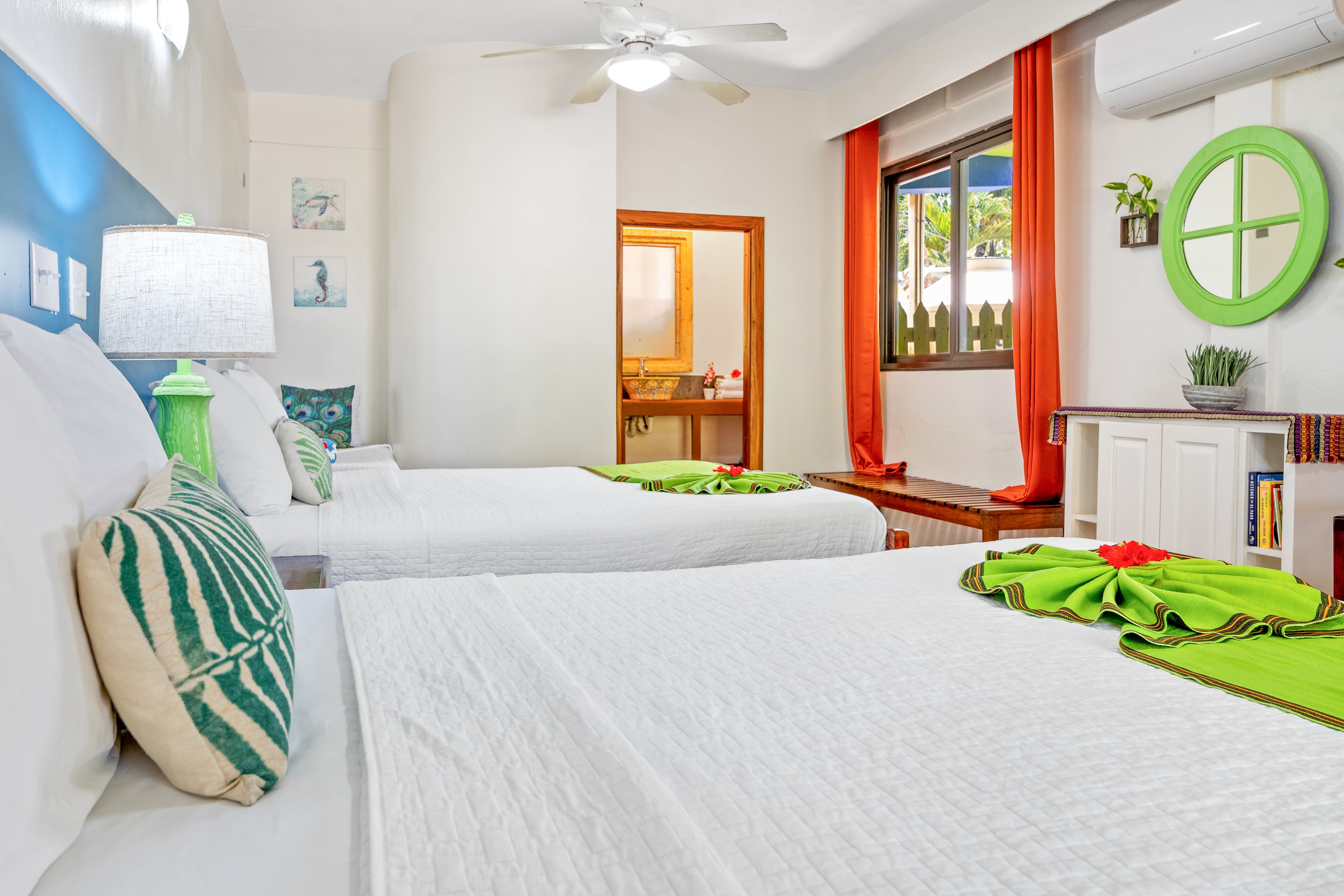 A brightly lit bedroom featuring two queen-size beds with white linens, lime green decorative runners, and patterned accent pillows. The room is decorated with vibrant orange curtains, a lime green circular wall mirror, and a small white shelving unit filled with books, while a door in the background opens into an adjacent bathroom.