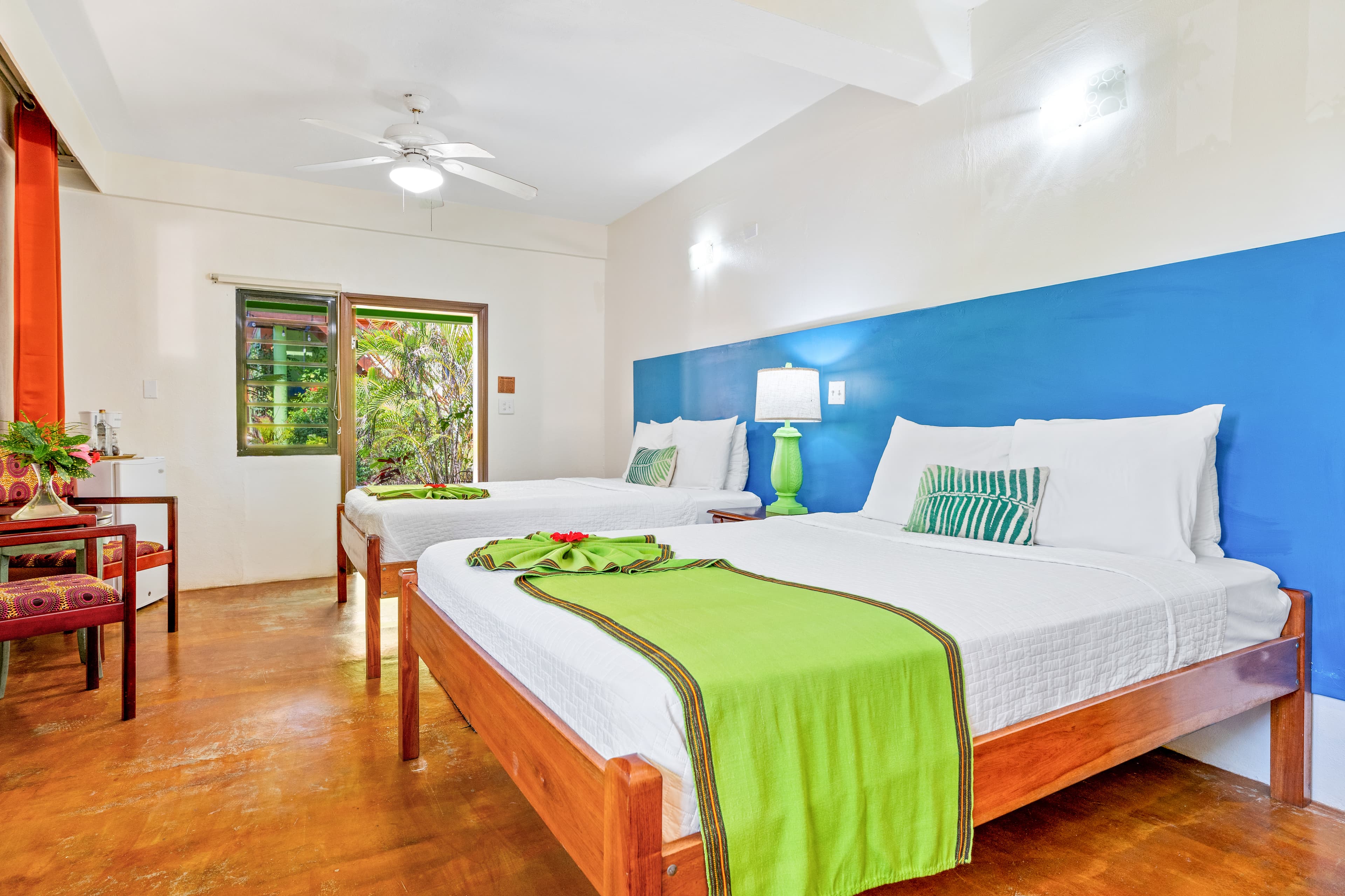 A brightly lit bedroom featuring two queen-size beds with white linens, vibrant lime-green runners, and patterned accent pillows. The room includes terracotta tile flooring, a blue accent wall behind the beds, and a glass door that opens to a lush outdoor area.