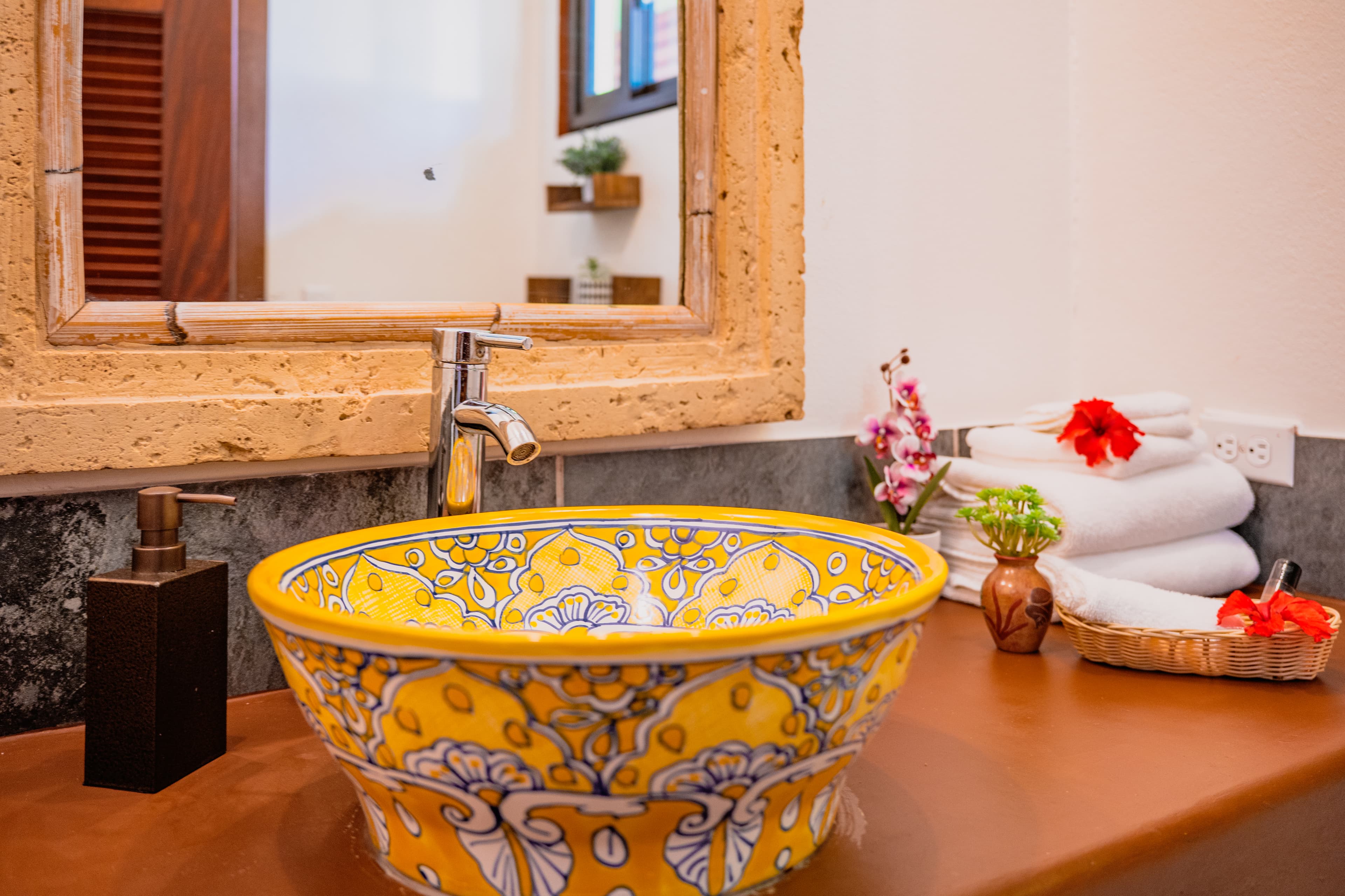 A vibrant yellow and blue patterned vessel sink sits on a smooth brown countertop in front of a mirror with a decorative, textured beige frame. The bathroom vanity is styled with a dark soap dispenser, a small wooden basket of toiletries, and a stack of clean white towels adorned with a bright red flower.