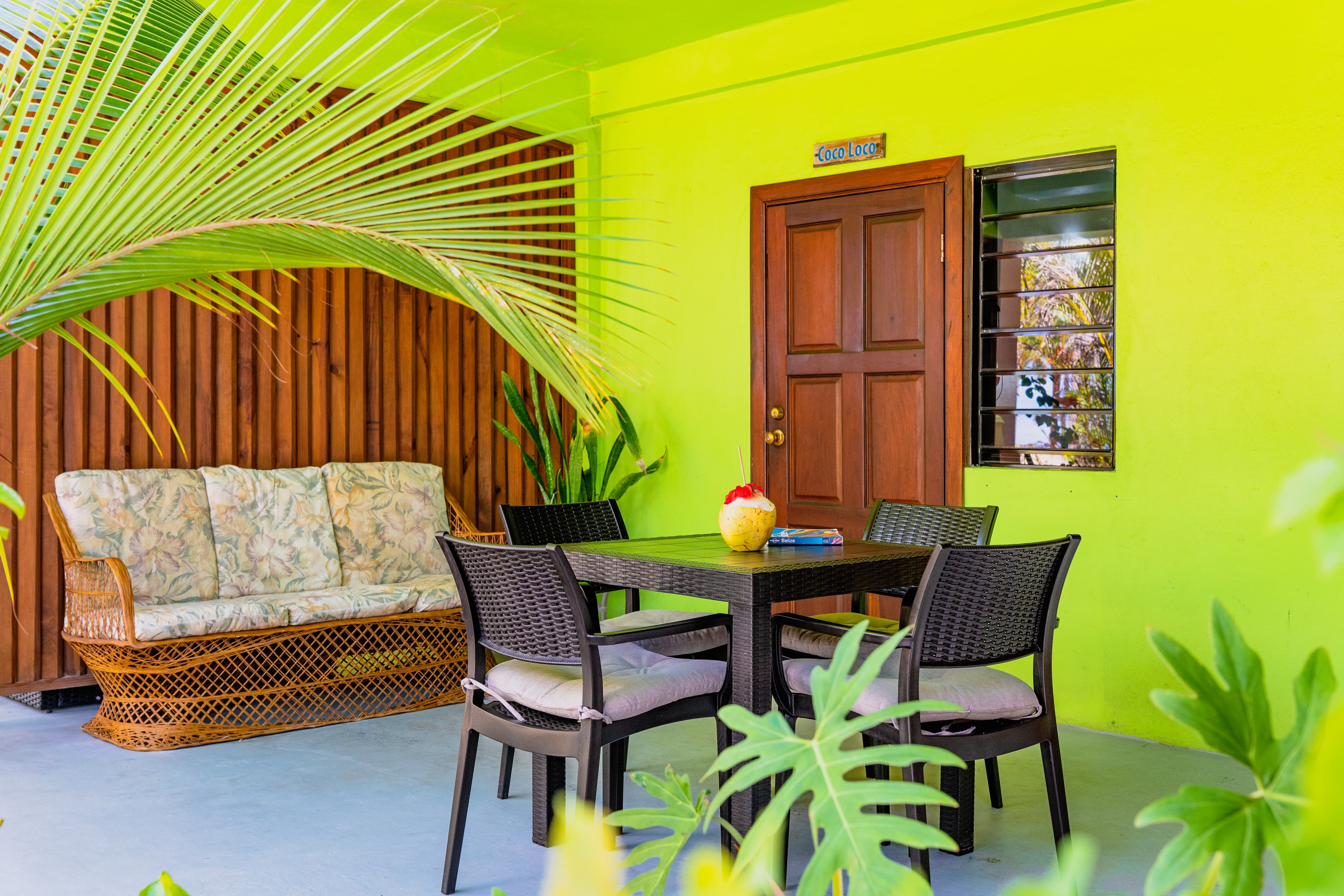 An outdoor patio with a lime-green wall and a grey floor. The area is furnished with a dark wood dining table and four chairs, as well as a wicker sofa with patterned cushions set against a wooden slat privacy screen. Large tropical green leaves frame the foreground.