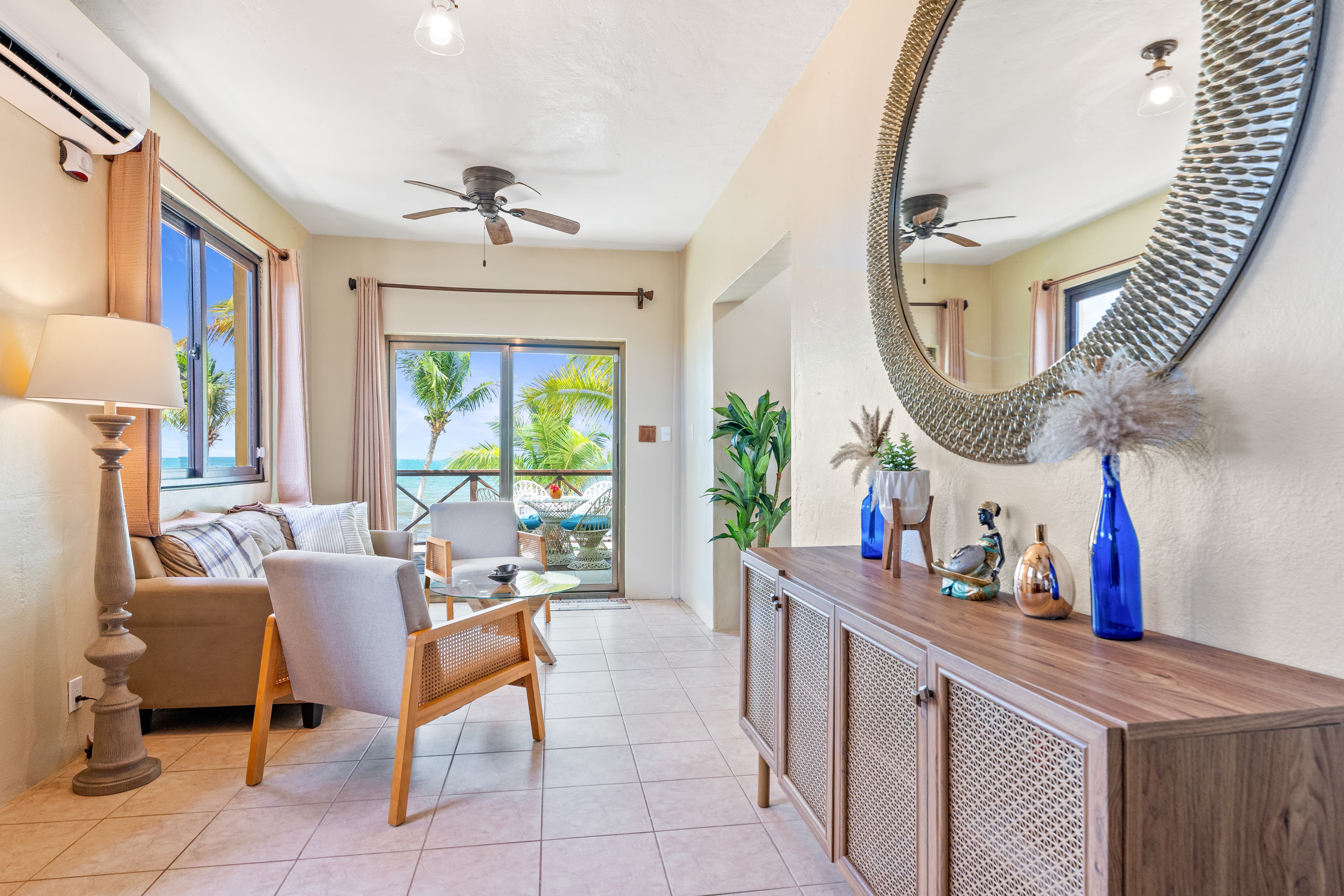 A bright living area featuring a tan sofa and two grey armchairs arranged around a glass coffee table. The room includes a long wooden sideboard under a large, round decorative mirror, a tall floor lamp, and a sliding glass door that opens to a patio with a view of tropical palm trees and the sea.