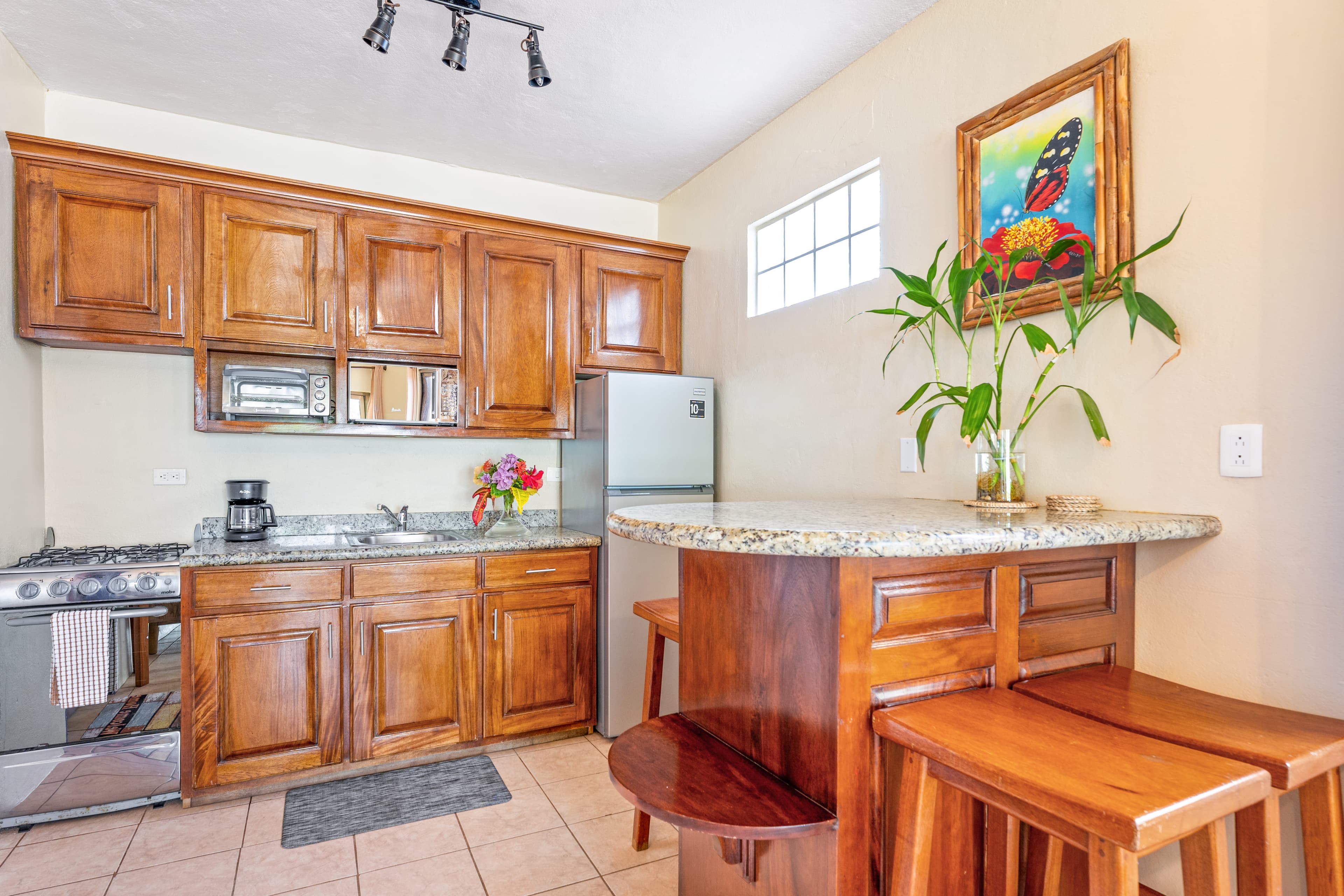 A small kitchen area with warm wood cabinetry and granite countertops, featuring a compact gas stove, microwave, and refrigerator. A tall wooden breakfast bar with matching stools is situated to the right, decorated with a vibrant tropical painting and a glass vase with green plant cuttings.