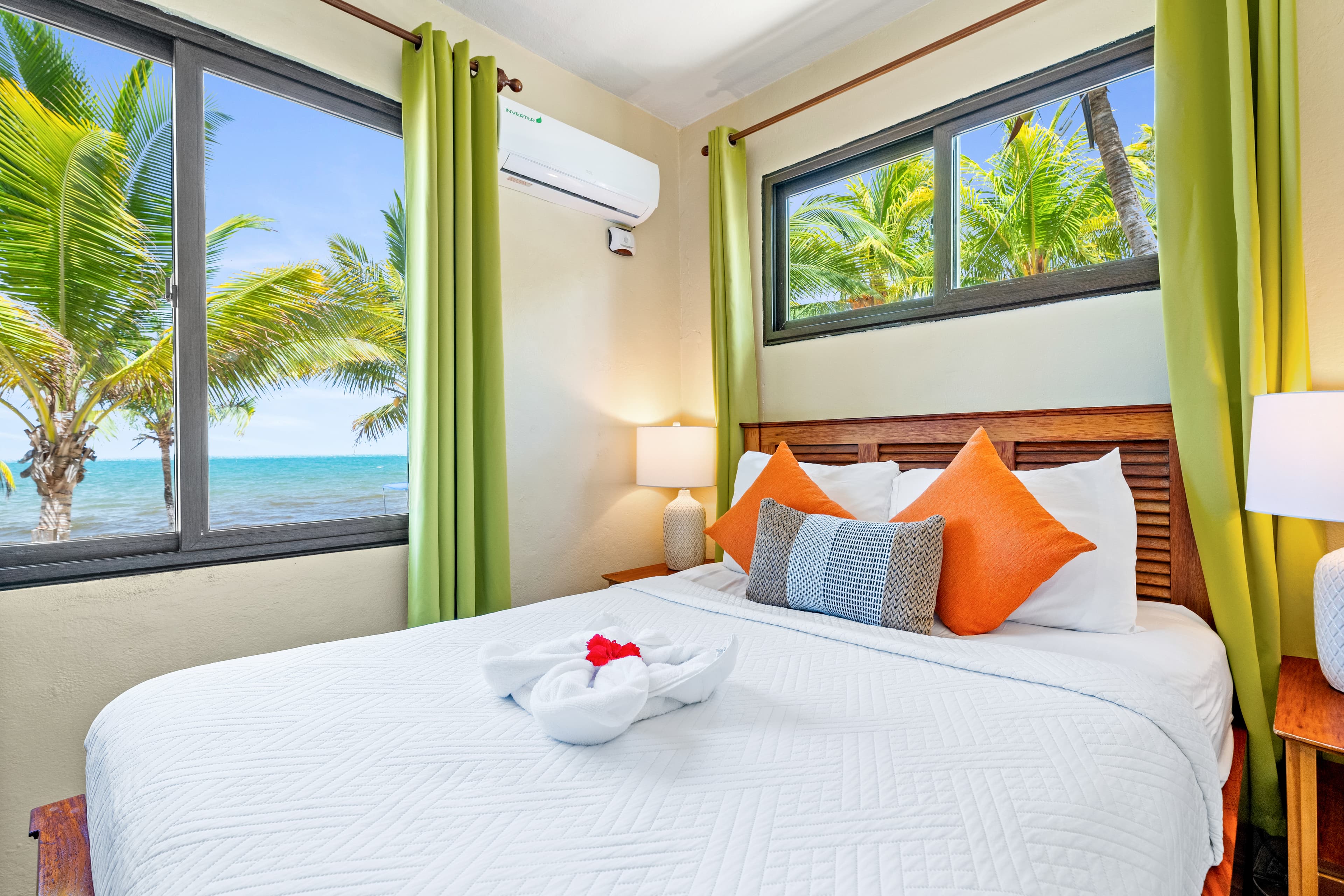 A bedroom with white walls and terracotta tile flooring featuring a queen-size bed with white linens, bright orange accent pillows, and a decorative white towel shaped like a flower. Two large windows with lime green curtains provide a view of lush palm trees and the turquoise sea.