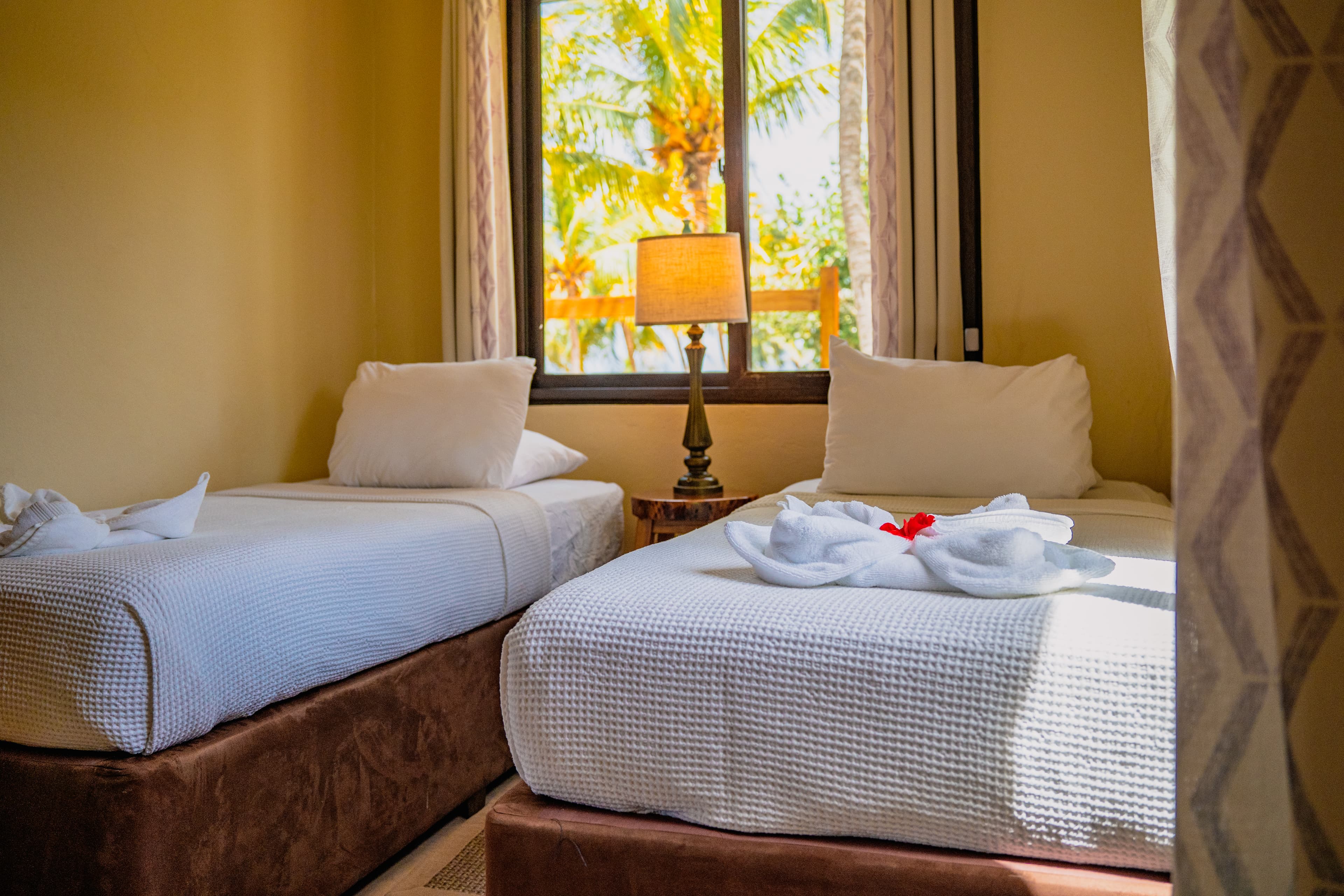 A brightly lit bedroom featuring two twin beds with brown bases and white linens, each adorned with a decorative white towel shaped like a flower. A small wooden nightstand with a classic lamp sits between the beds in front of a window that looks out onto lush green palm trees.