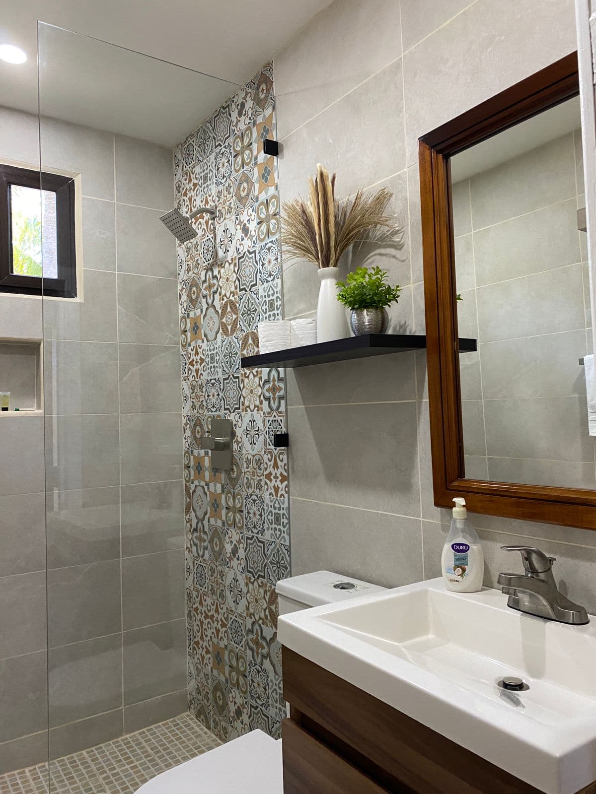 A modern bathroom featuring a white vessel sink on a wood-grain vanity with a dark wood-framed mirror. The space includes a white toilet and a walk-in shower with a glass partition and a decorative mosaic-tiled accent wall. Above the toilet, a black shelf holds a white vase with dried pampas grass and a small potted plant.