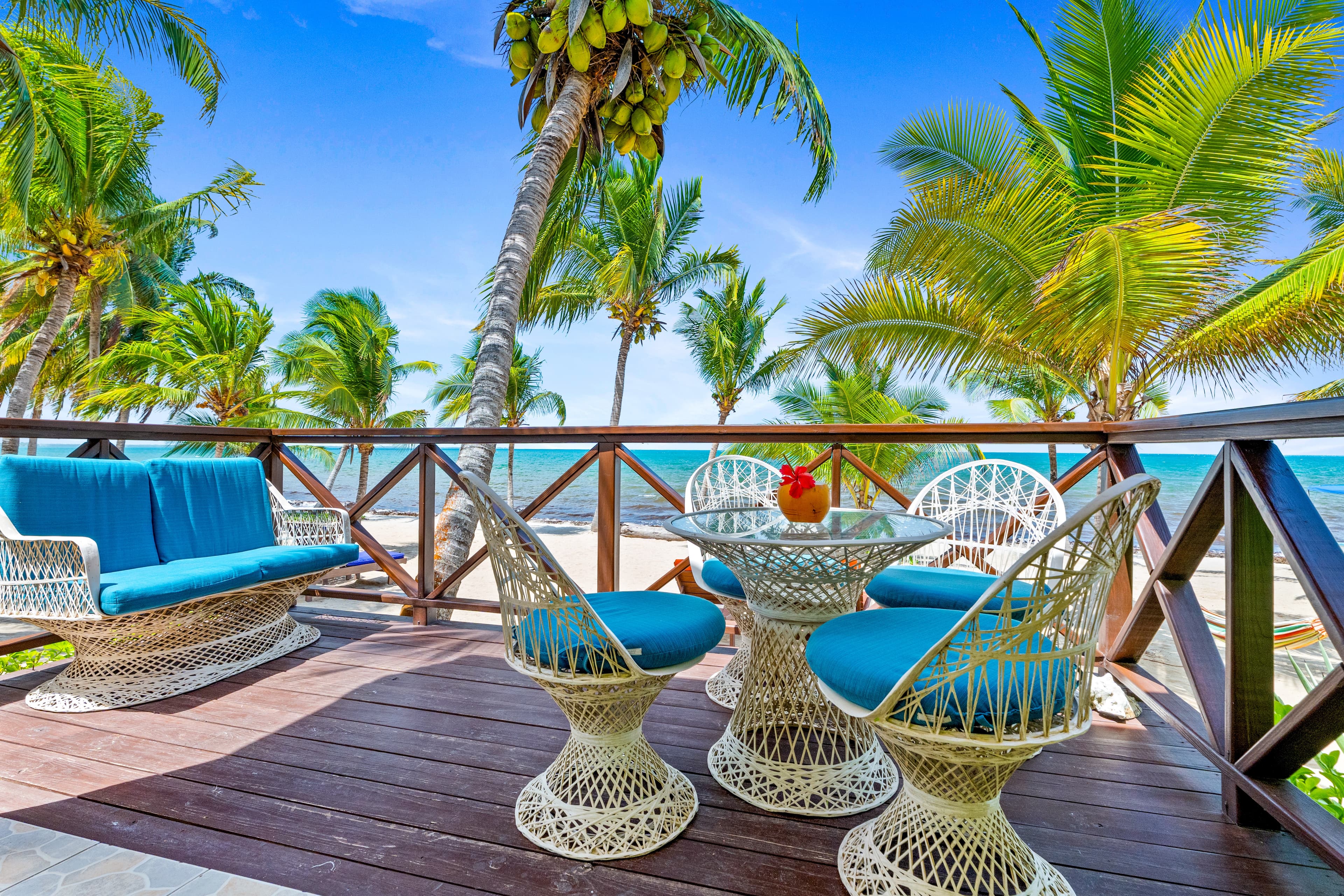 An outdoor wooden deck overlooks the turquoise sea and a sandy beach lined with palm trees under a clear blue sky. The space is furnished with white wicker seating featuring bright blue cushions, including a sofa and a circular dining set centered around a glass-topped table.