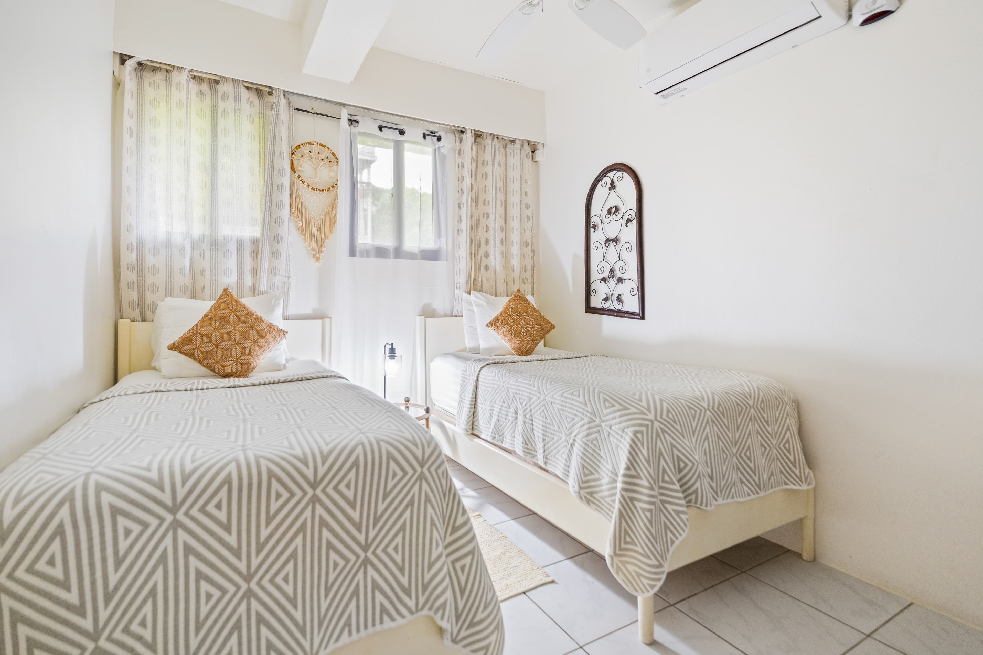 A bright, minimalist bedroom featuring two twin beds with white and grey geometric patterned linens and ochre accent pillows. The room has white walls, light-colored tile flooring, and a large window with patterned curtains, while a decorative dark-framed wall hanging adds a classic touch to the space.