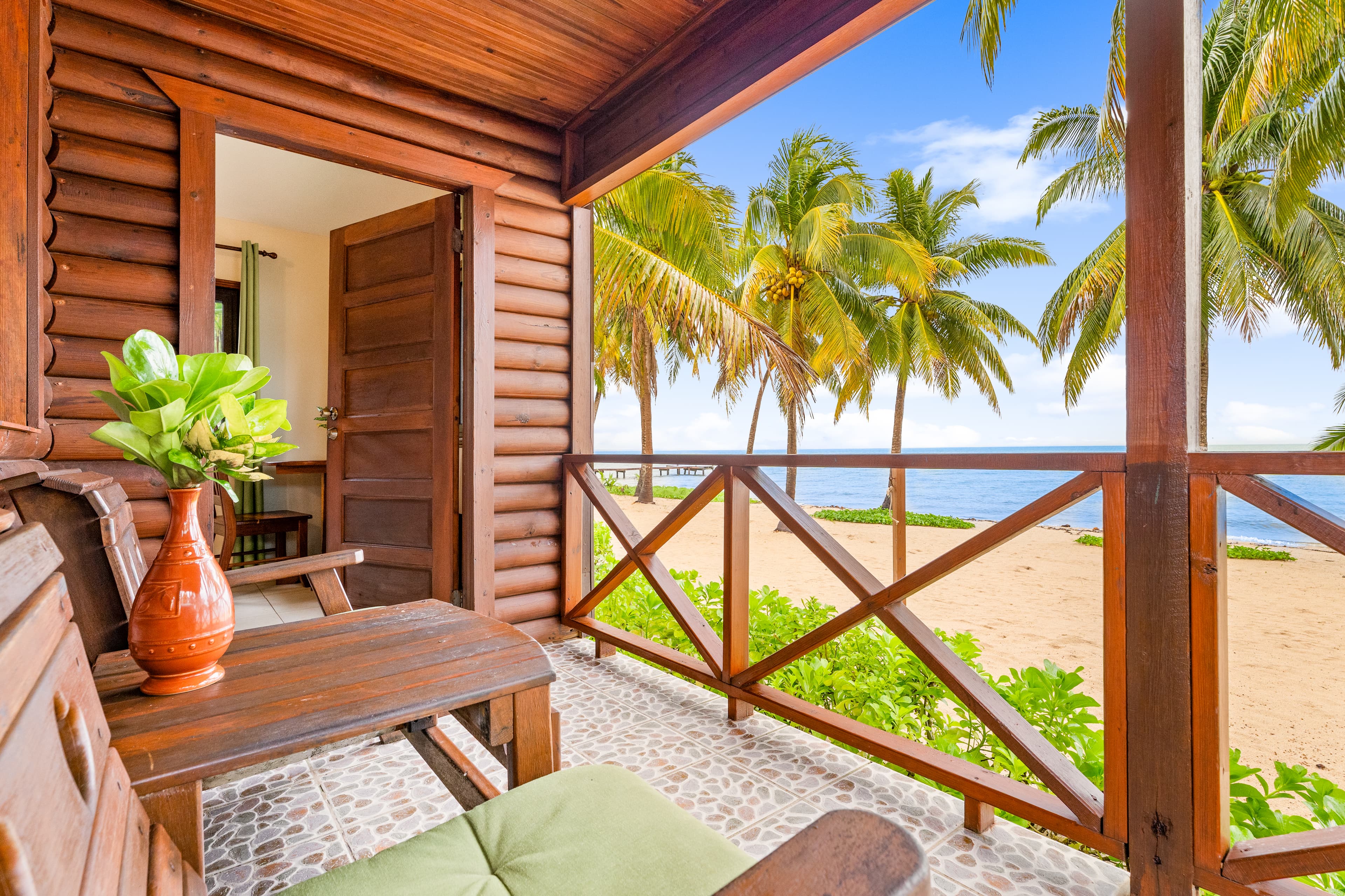 A narrow wooden balcony and railing overlooks a sandy beach lined with palm trees and a calm, turquoise ocean under a bright blue sky. The balcony is furnished with wooden chairs featuring green cushions and a small side table topped with a vibrant orange vase holding green tropical foliage.