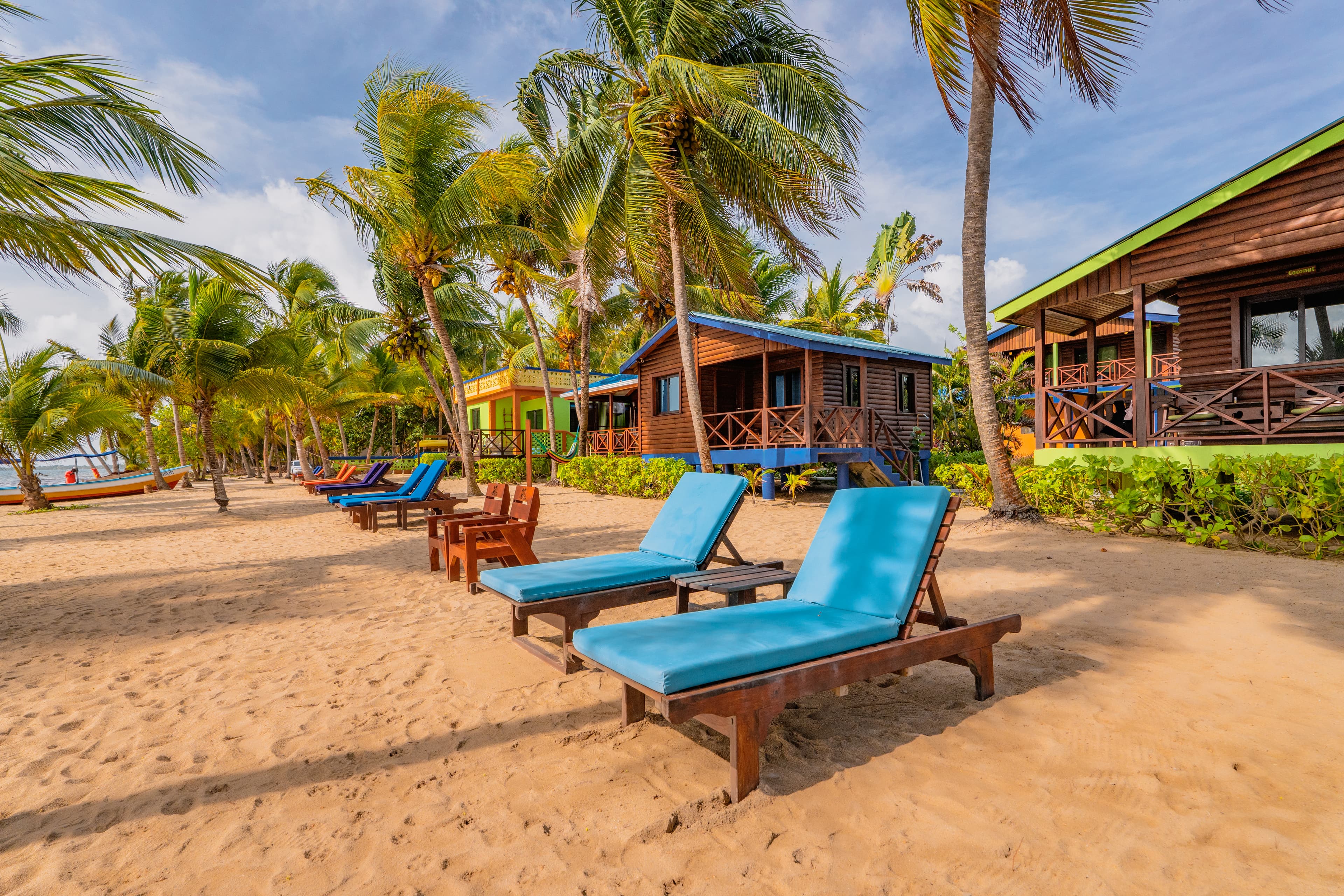 Several wooden beachfront bungalows with dark roofs sit nestled among tall, leaning palm trees on a sandy beach. Bright blue lounge chairs are arranged in a row along the sand in front of the cabins, all under a blue sky with soft white clouds.