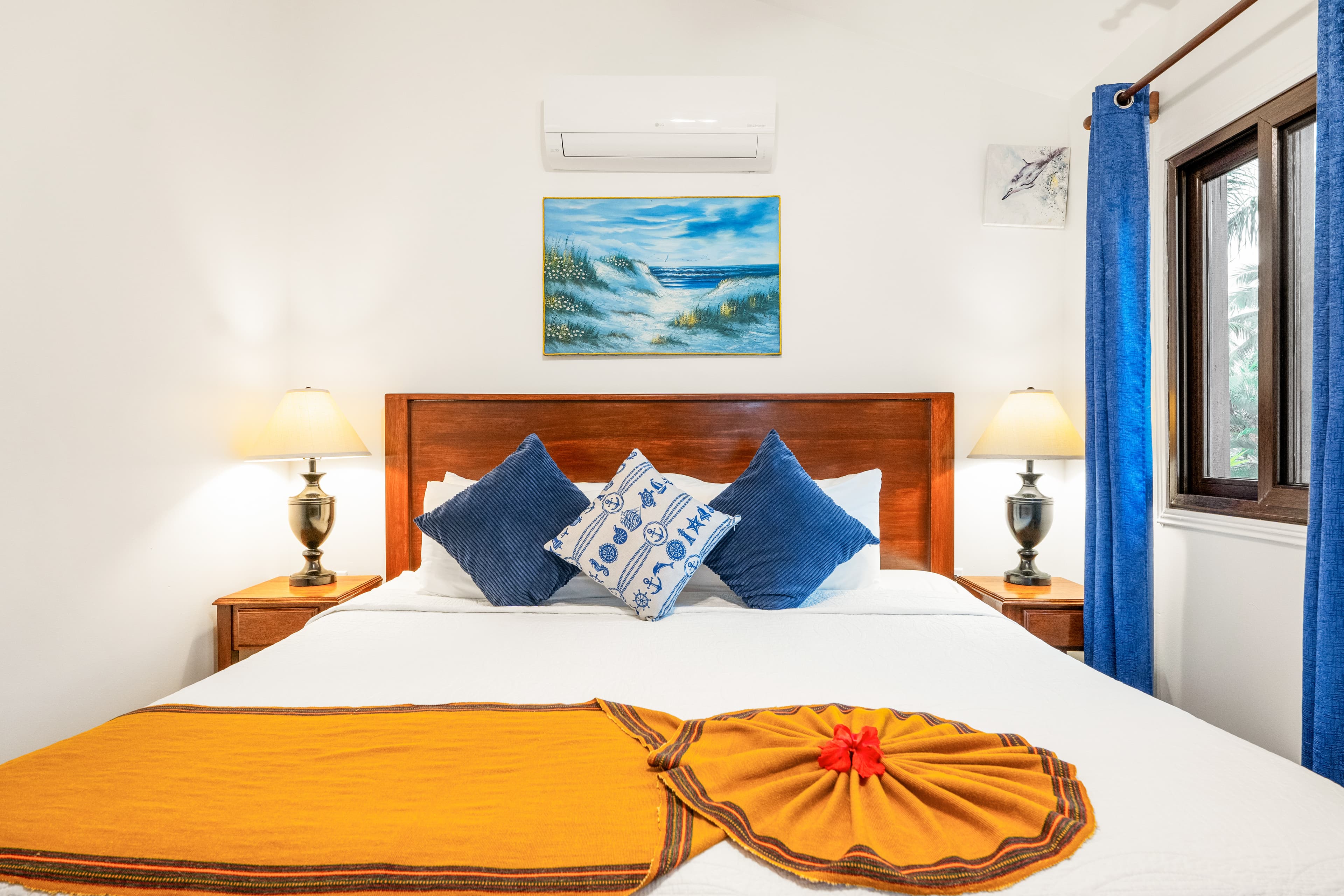 A brightly lit bedroom featuring a king-size bed with white linens, a vibrant golden-orange runner, and a white towel folded into a decorative flower with a red blossom in the center. The bed is styled with two solid blue pillows and a central patterned accent pillow, flanked by two wooden nightstands with classic lamps, while a painting of ocean waves and bright blue curtains complete the tropical aesthetic.