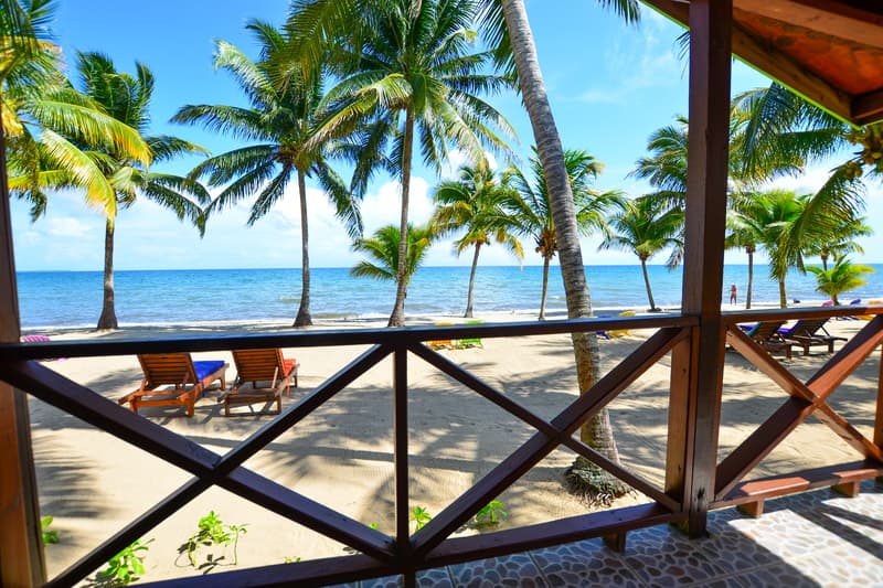 A view from an outdoor wooden balcony overlooks a sandy beach dotted with palm trees and sun loungers. The turquoise sea stretches to the horizon under a bright, clear blue sky.