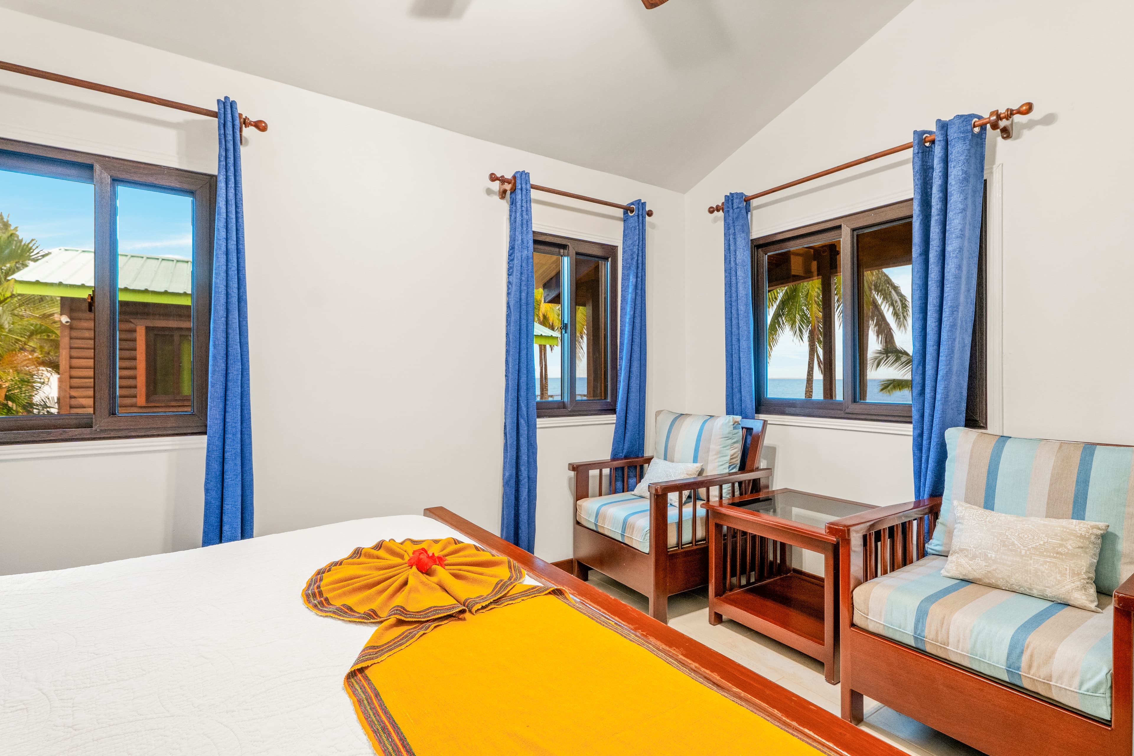 A brightly lit bedroom featuring a king bed with white linens, a vibrant golden-orange runner, and a white towel folded into a decorative flower with a red blossom in the center. Two wooden armchairs with blue and tan striped cushions are positioned in front of three windows framed by bright blue curtains, offering a view of a lush tropical beach.