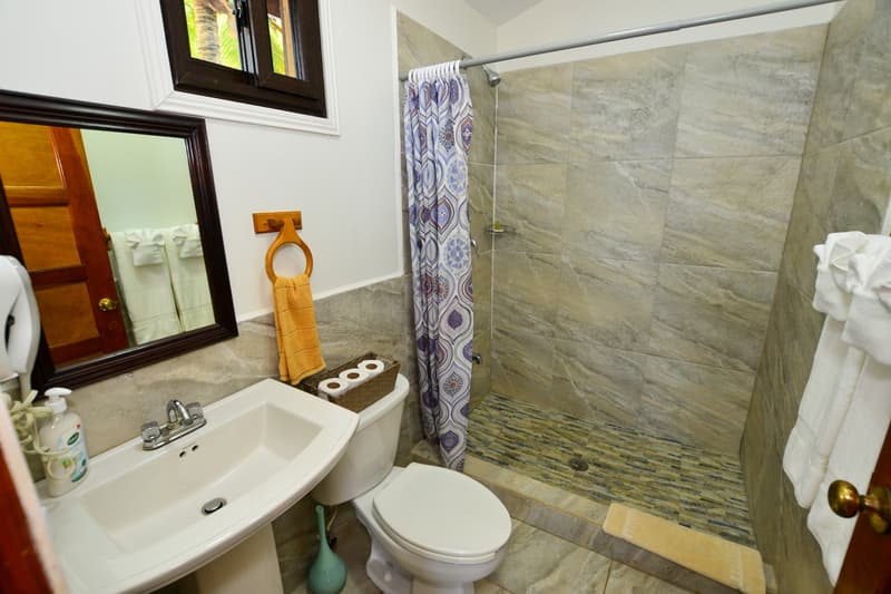 An indoor bathroom with stone-patterned wall tiles and a walk-in shower featuring a patterned blue and white curtain. The room includes a white pedestal sink and toilet, a dark wood-framed mirror, and a stack of white towels on the right, with a small window at the top left showing tropical greenery.