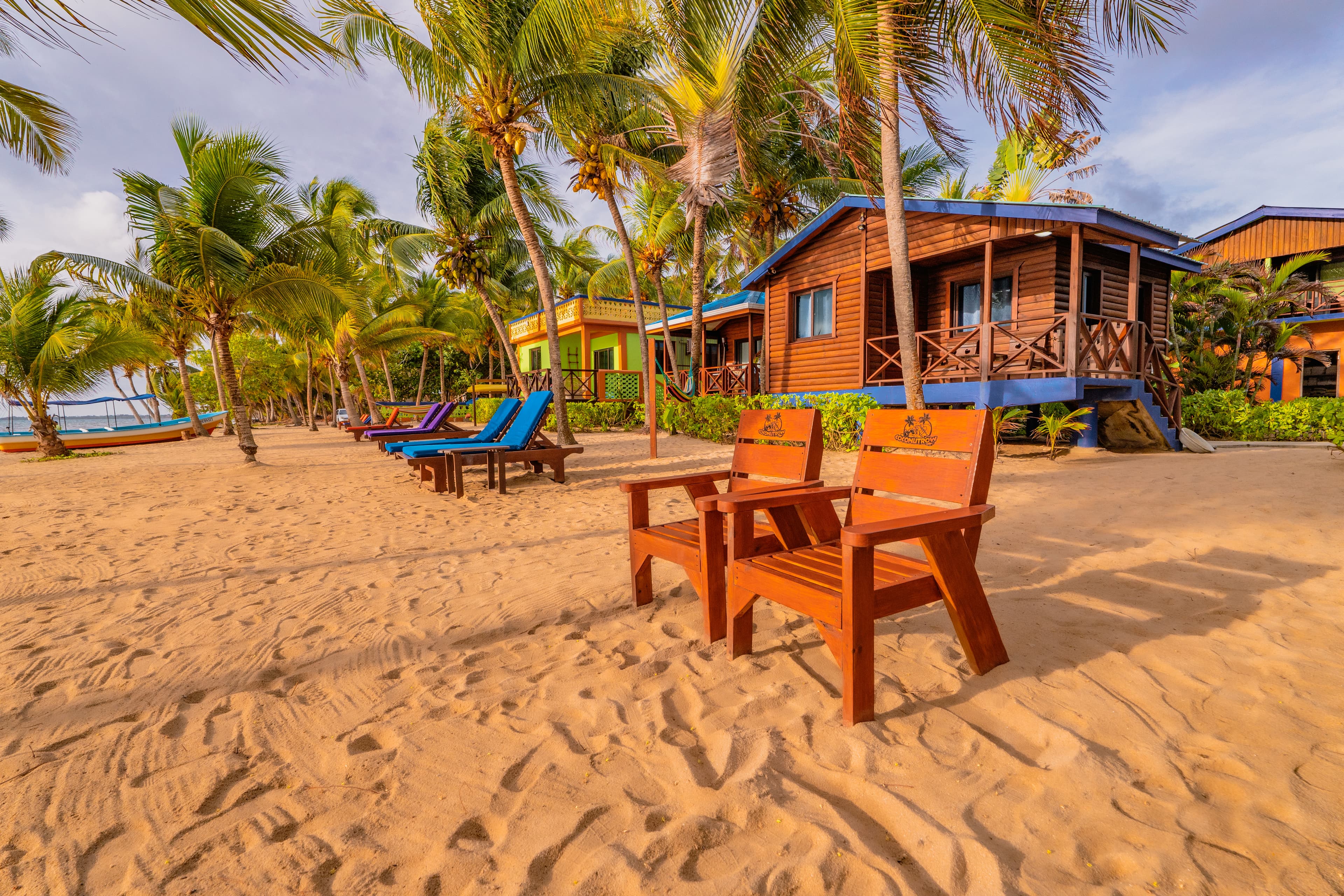 A row of wooden beachfront bungalows with dark roofs nestled among leaning palm trees on a sandy beach. Bright blue lounge chairs and wooden Adirondack chairs are arranged on the sand, all under a blue sky with soft, wispy clouds.