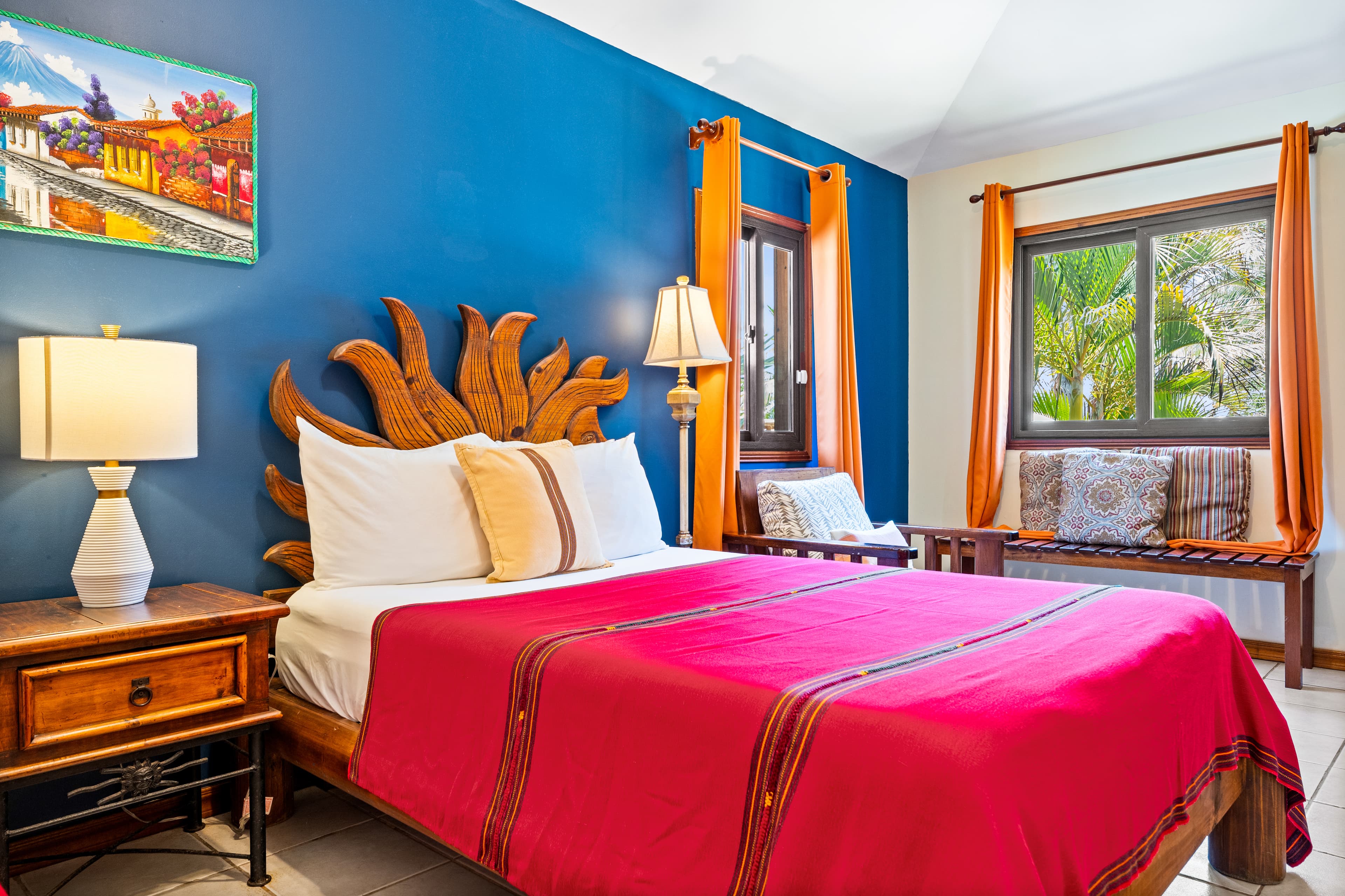 A vibrant bedroom featuring a full-size bed with a bright red cover and a unique, sunburst-shaped wooden headboard set against a deep blue accent wall. The room includes a wooden nightstand with a white lamp, an orange-curtained window, and a built-in bench with patterned cushions under a second window that overlooks lush tropical greenery.