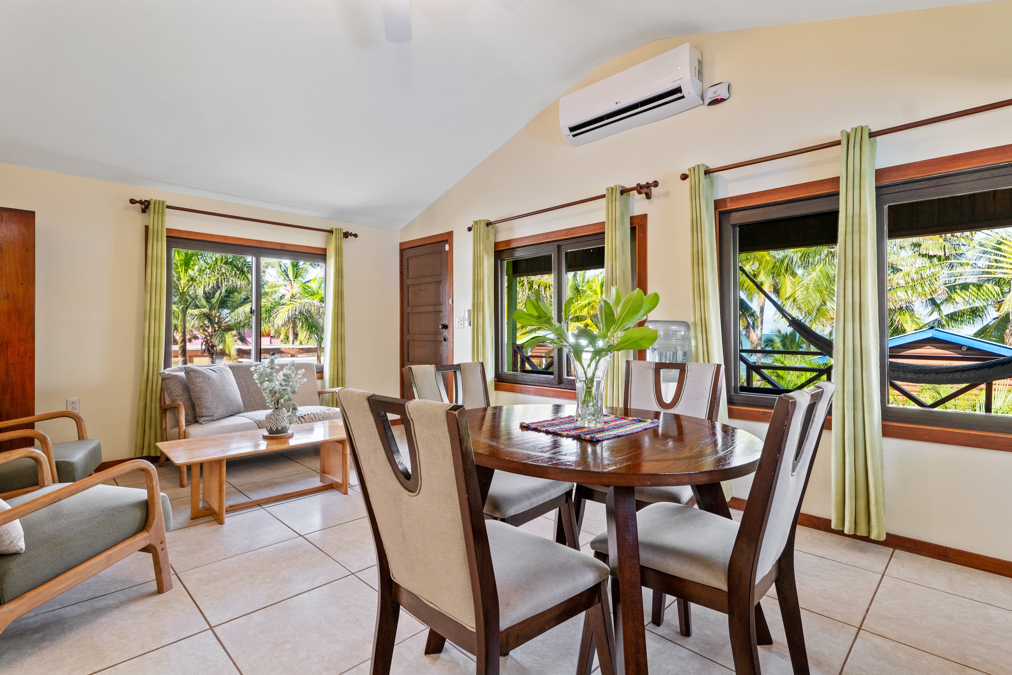 An open-concept living and dining area with light-colored tile flooring and a vaulted ceiling. A dark wood oval dining table with four cushioned chairs sits in the foreground, while a seating area with a sofa, armchairs, and a coffee table is visible in the background. Multiple windows with lime-green curtains offer views of a lush tropical patio and a hammock.