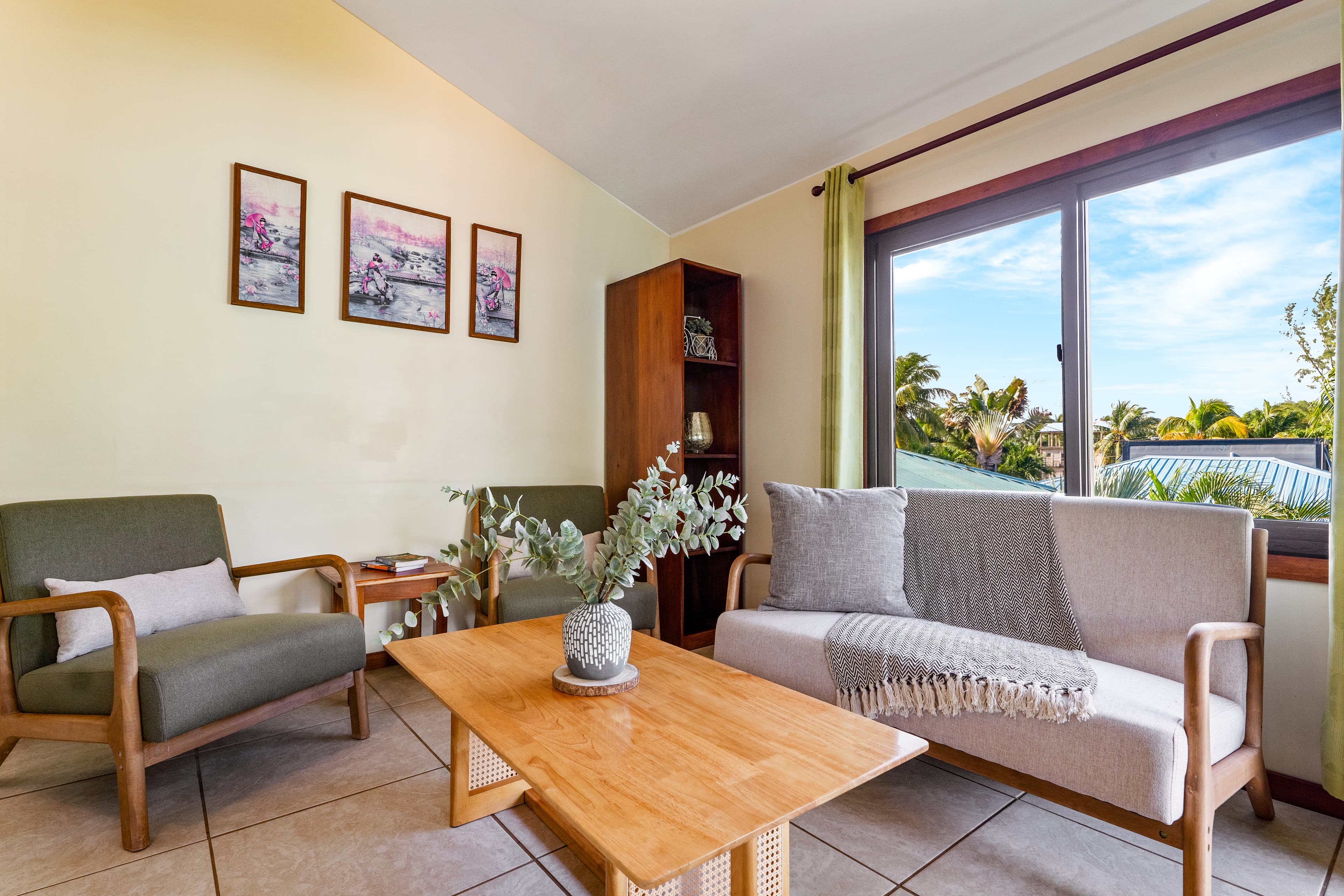 A spacious, light-filled living and dining area with a vaulted ceiling and beige walls. In the foreground, a dark wood oval dining table is set with four chairs that have beige upholstered backs. The background features a cozy seating area with a light grey sofa and two grey armchairs arranged around a wooden coffee table, while multiple windows framed with lime-green curtains look out onto a tropical property with palm trees.
