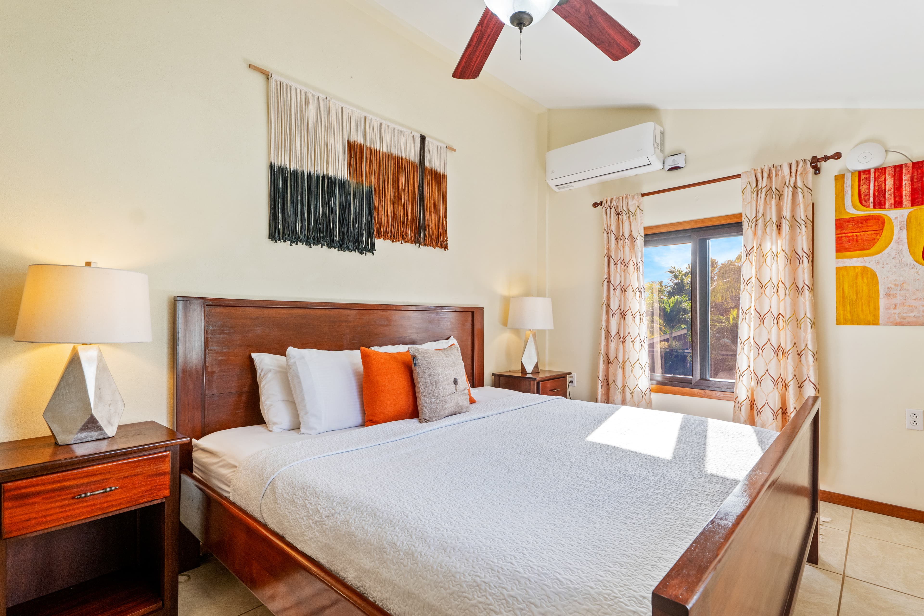 A brightly lit bedroom featuring a dark wood king-size bed with white linens and a vibrant orange accent pillow. The room has neutral beige walls decorated with a large woven textile wall hanging, two wooden nightstands with geometric lamps, and a window with patterned orange and white curtains that let in natural light.