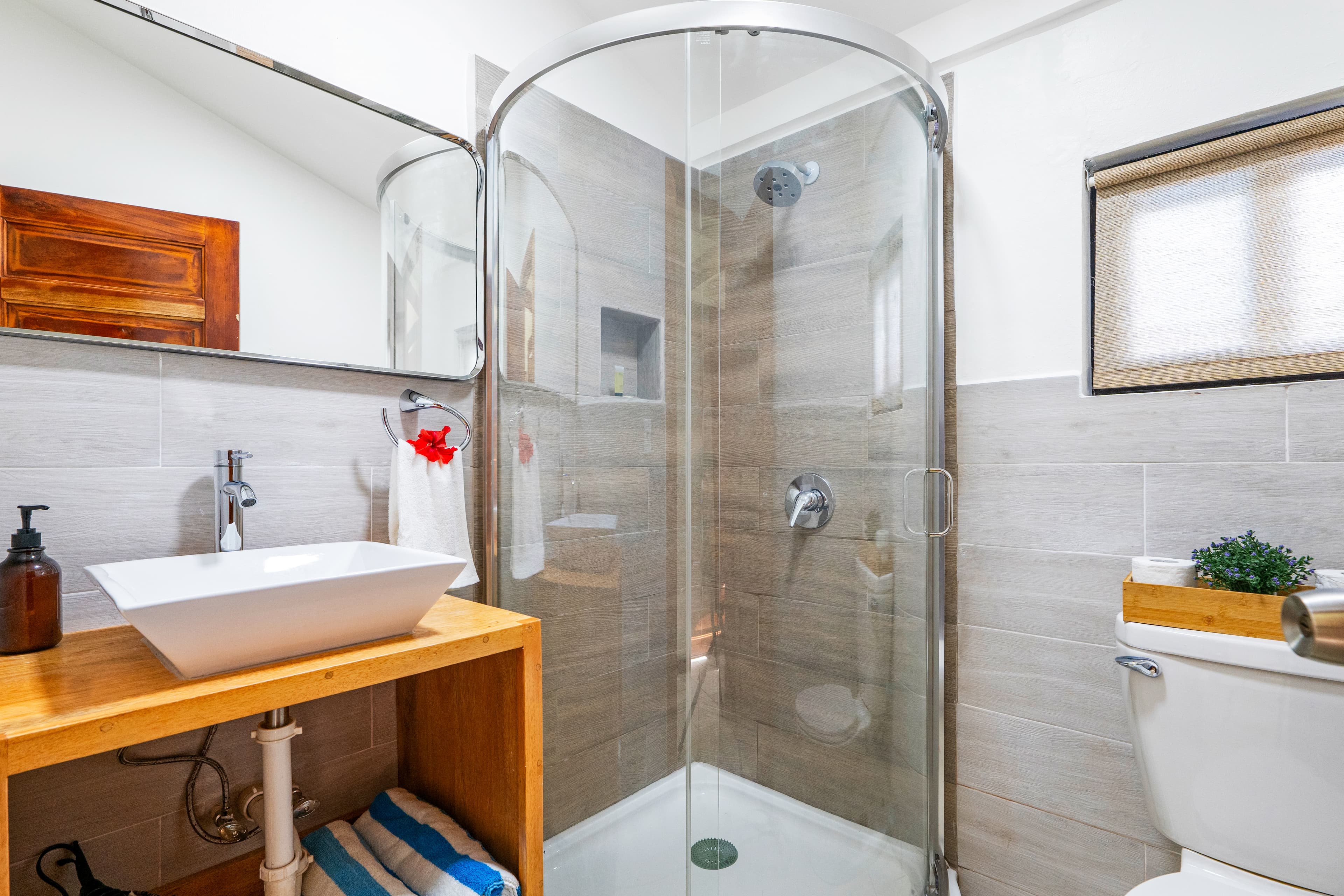 A clean, modern bathroom with grey wall tiles and a walk-in shower featuring a rounded glass enclosure and wood-look tile accents. The space includes a white square vessel sink on a light wood vanity, a large frameless mirror, and a white toilet decorated with a small potted plant in a wooden box.