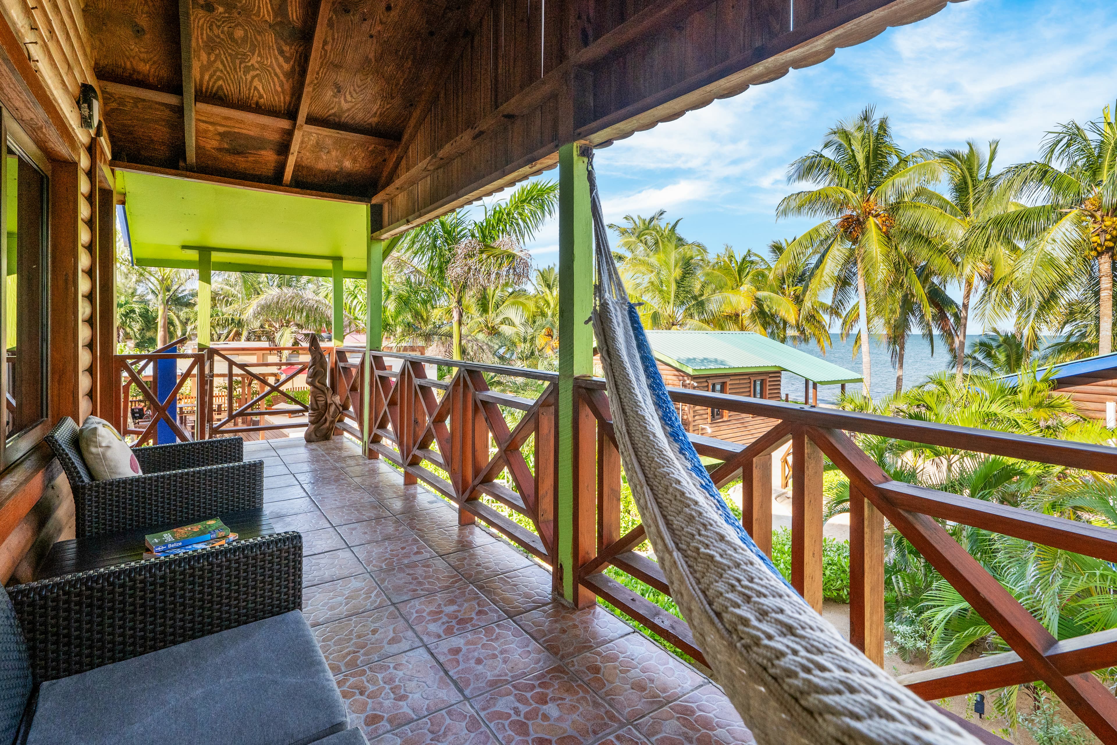 A spacious wooden balcony with a vaulted ceiling and tiled floor features a seating area with dark wicker armchairs and a white woven hammock. The balcony is enclosed by a dark wood railing and offers a scenic view of tropical greenery, palm trees, and the sea under a bright blue sky.