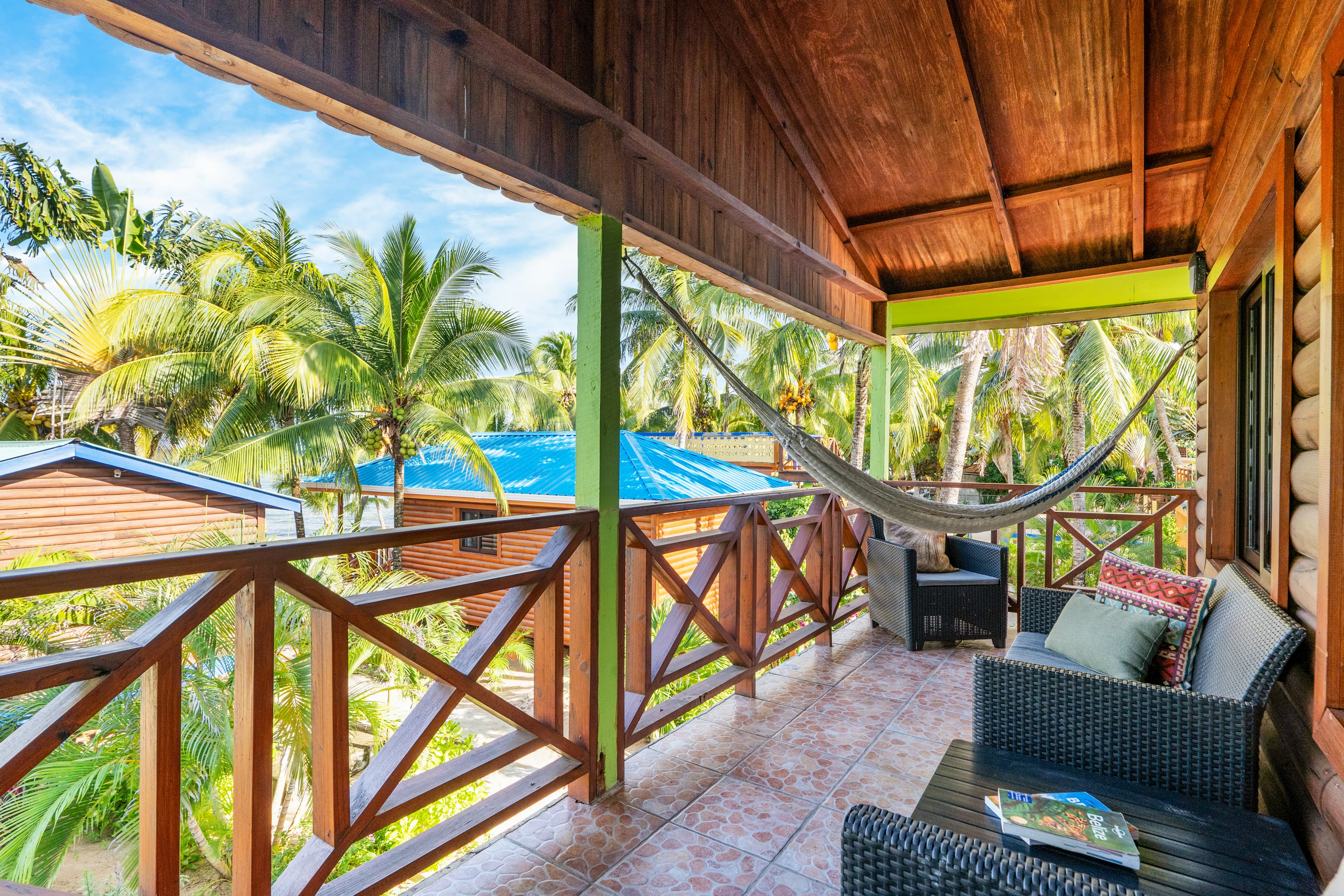 A spacious wooden balcony with a vaulted ceiling and tiled floor features a seating area with dark wicker armchairs and a white woven hammock. The balcony is enclosed by a dark wood railing and offers a scenic view of tropical greenery, palm trees, and the sea under a bright blue sky.