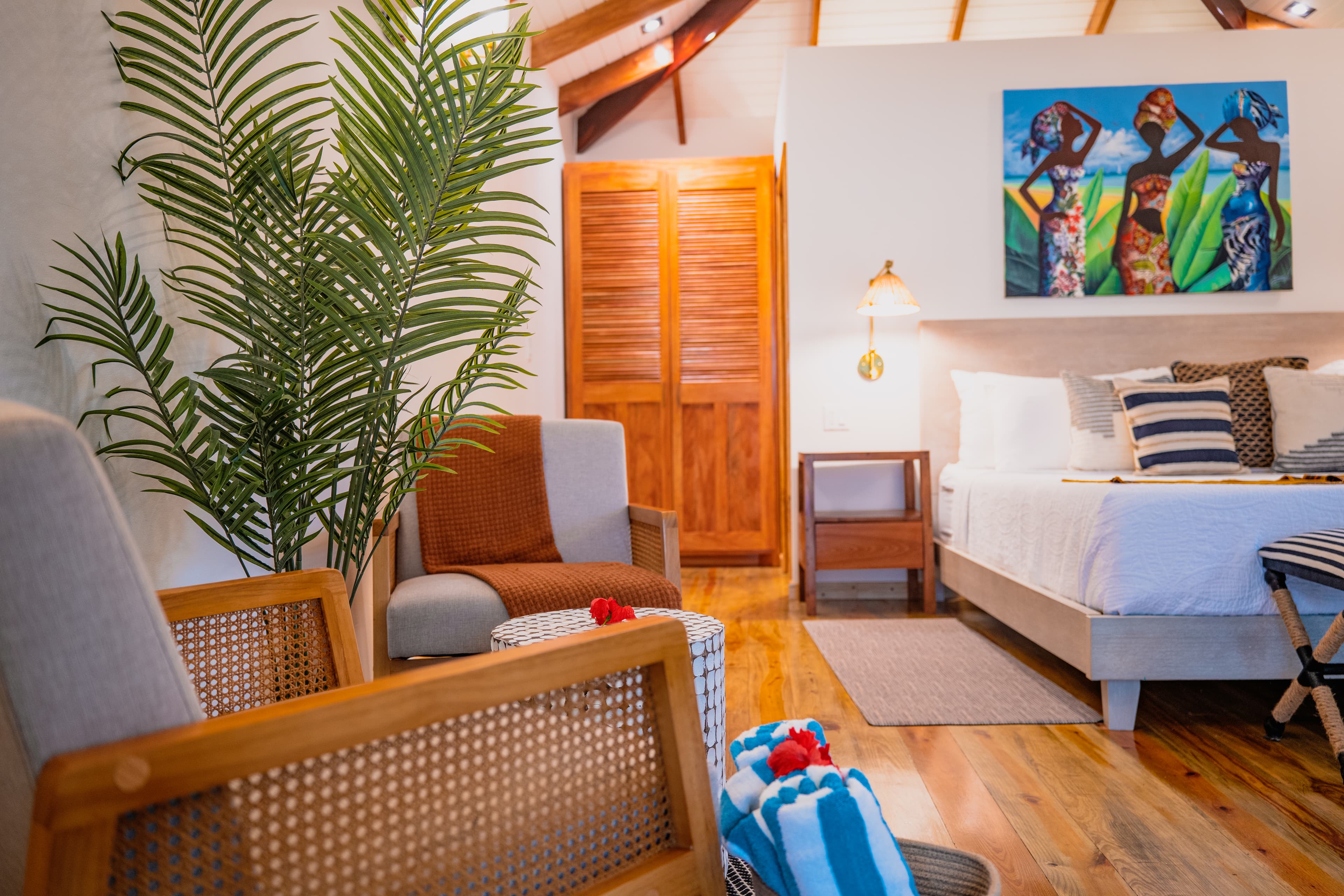 A bedroom with light wood flooring and a vaulted ceiling, featuring a seating area with a wooden armchair and a large potted palm plant in the foreground. In the background, a white bed is decorated with blue and white striped pillows and a colorful painting of three women hangs on the wall above the headboard.
