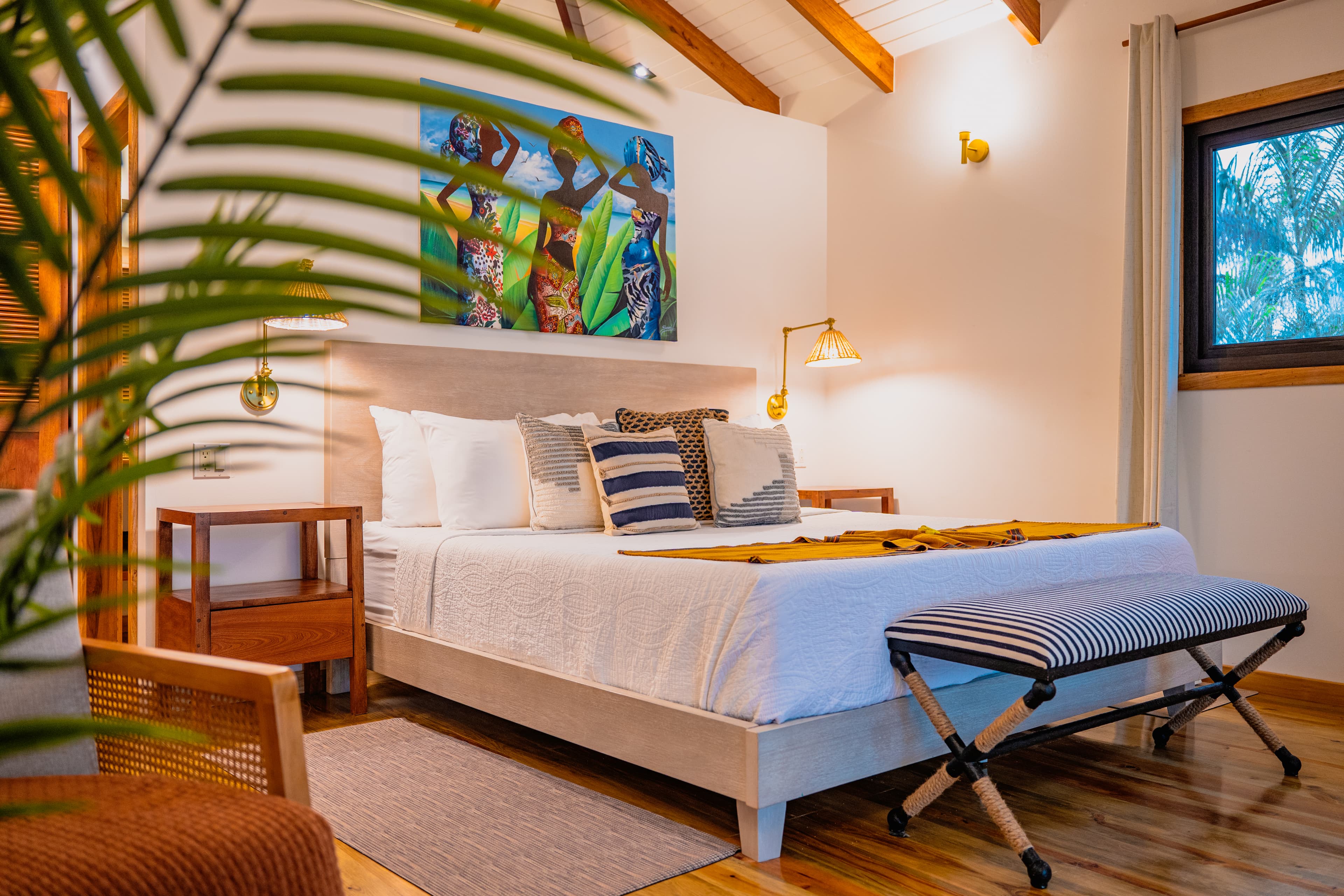 A bedroom with light wood flooring and a vaulted ceiling, featuring a seating area with a wooden armchair and a large potted palm plant in the foreground. In the background, a white bed is decorated with blue and white striped pillows, and a colorful painting of three women hangs on the wall above the headboard.