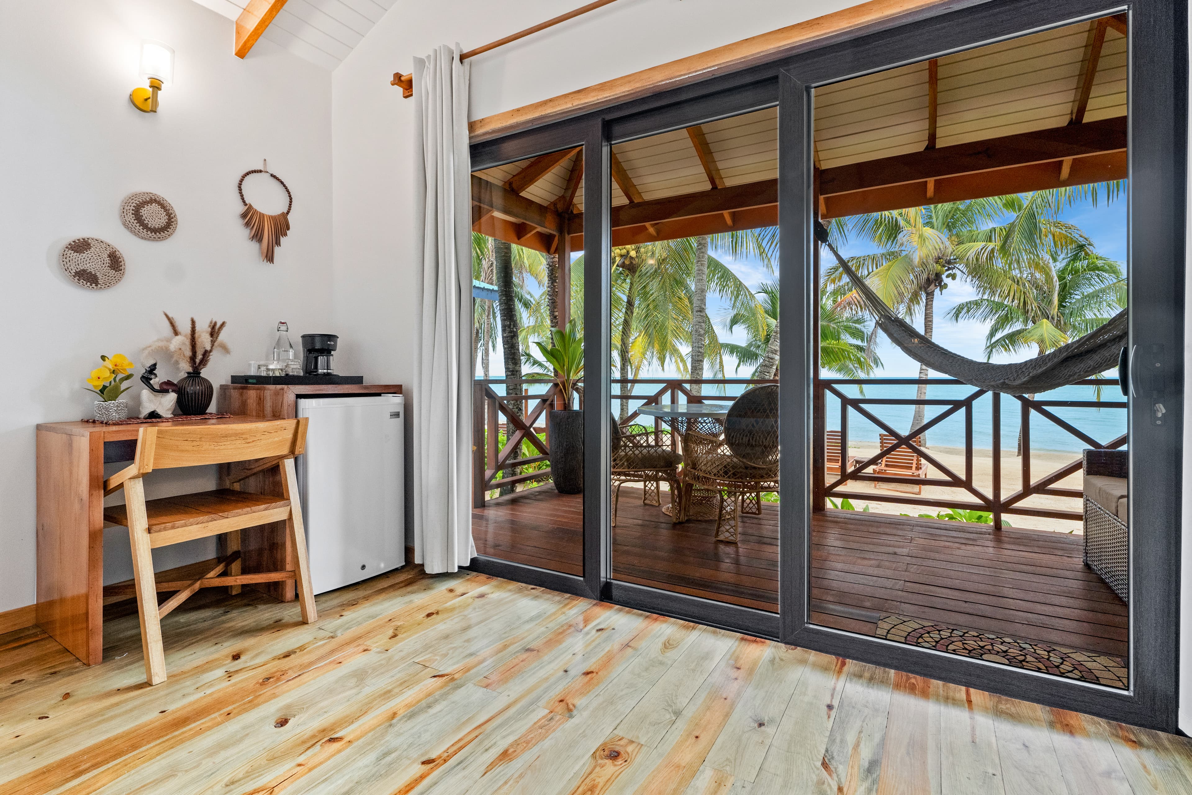 A bedroom with light wood flooring and a vaulted ceiling features a wooden desk and chair next to a small white refrigerator. Large sliding glass doors open onto a covered wooden balcony with a hammock, offering a view of a sandy beach, palm trees, and the ocean.