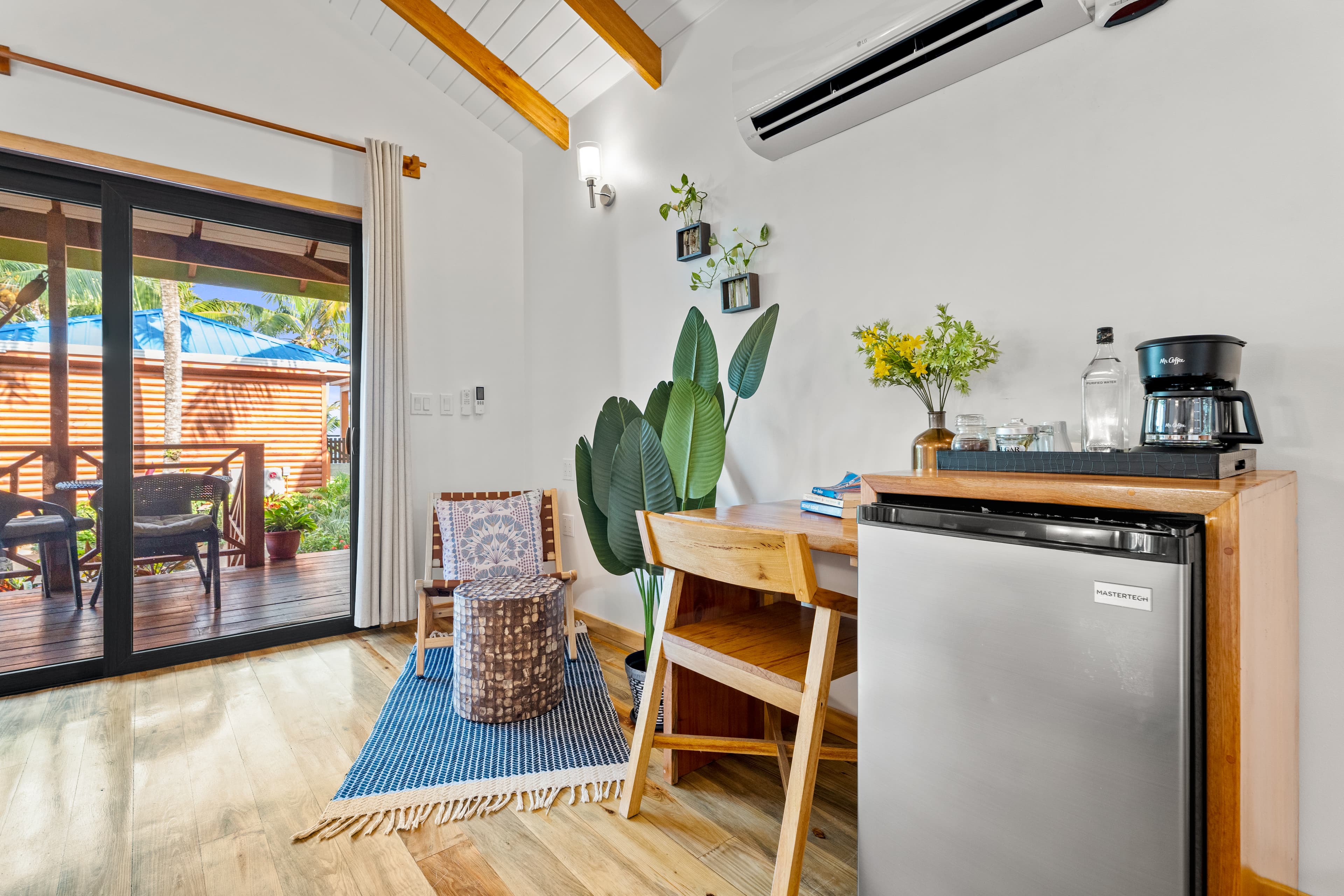 A bedroom with light-colored wooden floors and a vaulted wood-beam ceiling features a small wooden desk with a chair and a compact silver refrigerator. In the corner, a wooden armchair sits on a blue rug next to a large potted plant, while large sliding glass doors open onto a wooden balcony with a view of tropical greenery.