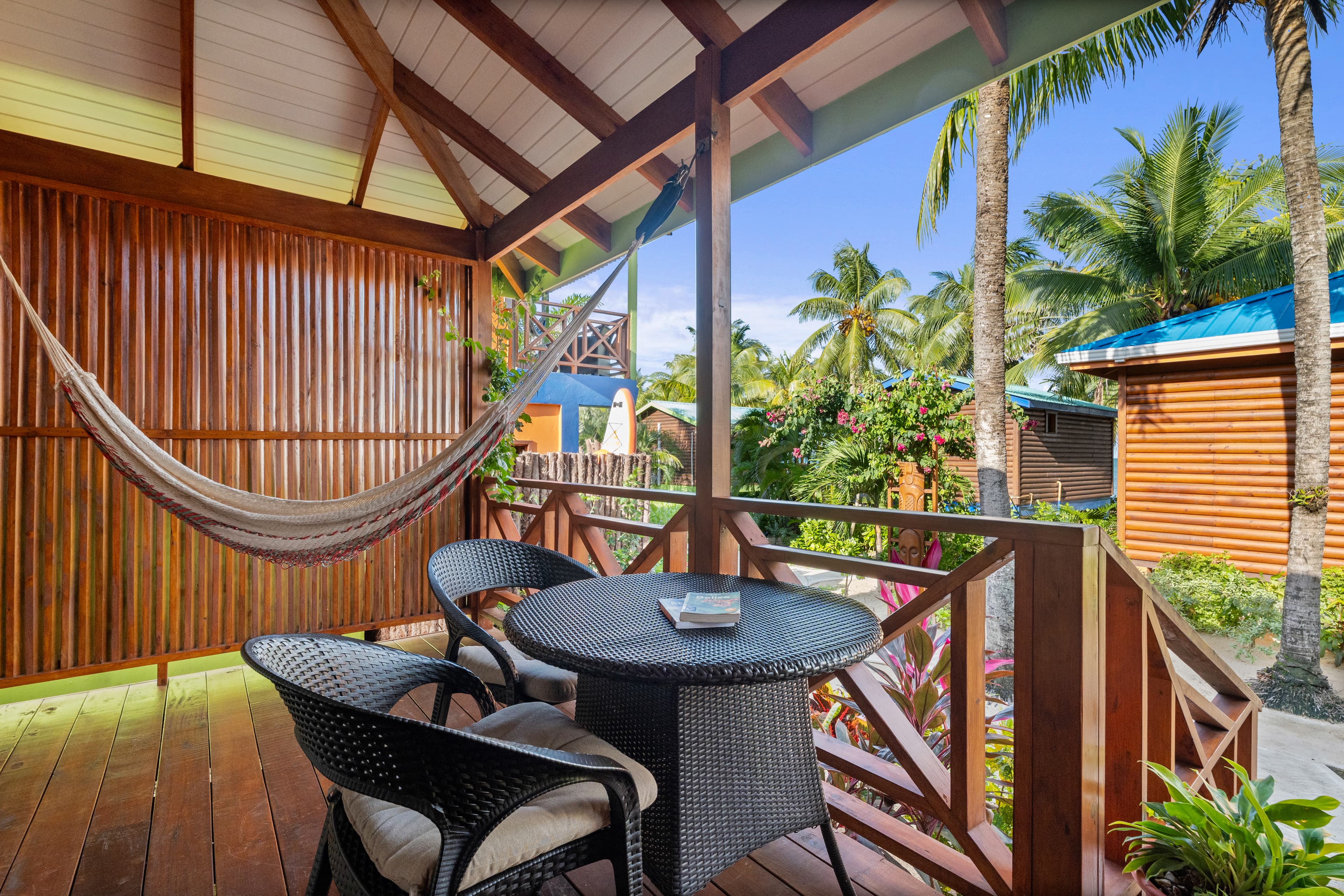 A spacious wooden balcony with a vaulted ceiling and dark wood railing features a small black table with two wicker armchairs and a striped hammock. The balcony offers a view of other wooden bungalows and lush tropical greenery under a clear blue sky.