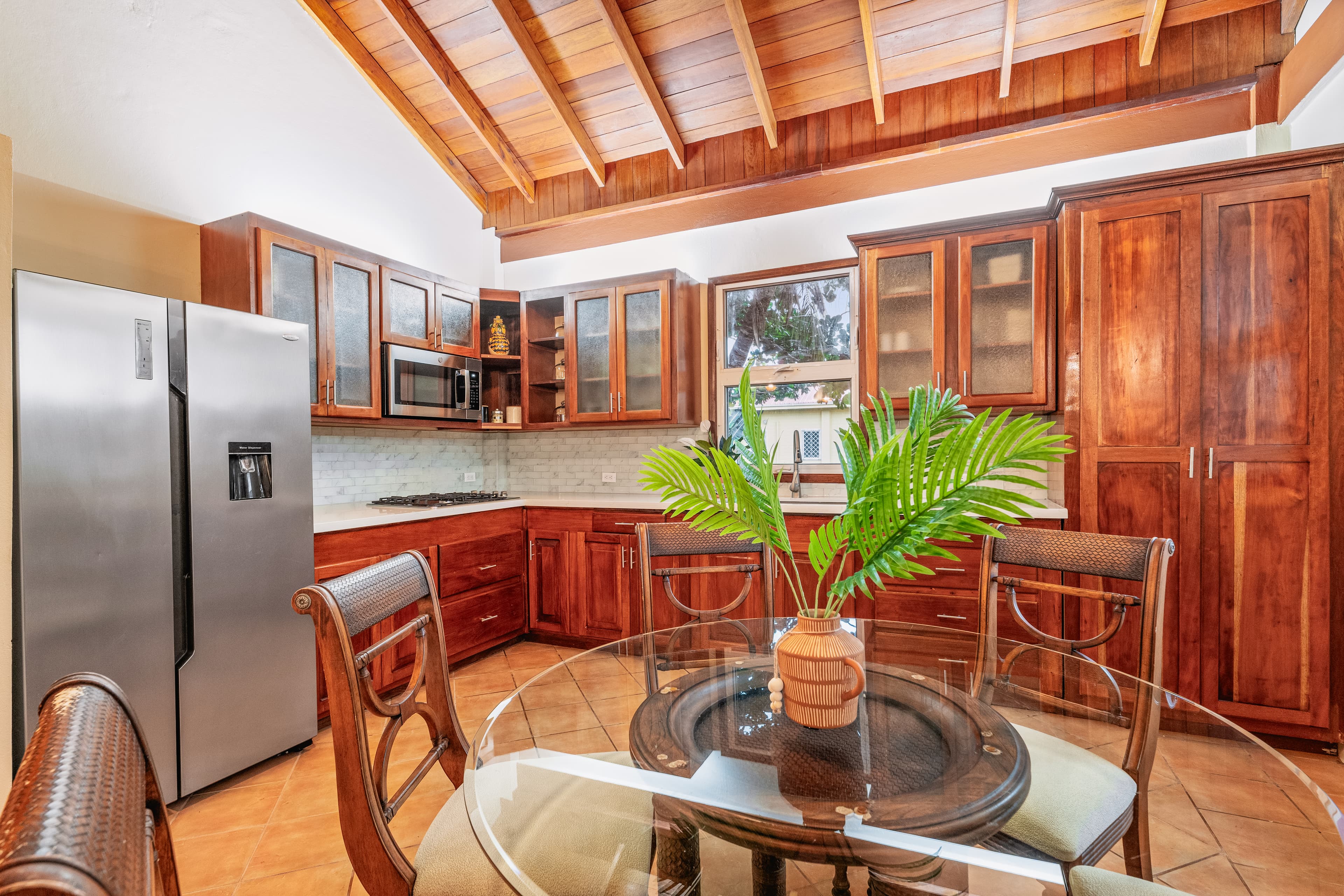 A spacious kitchen with a vaulted wooden ceiling and terracotta tile floors featuring rich wood cabinetry and a large stainless steel refrigerator. A glass-topped dining table with four wooden chairs sits in the foreground, decorated with a small plant in a brown vase.