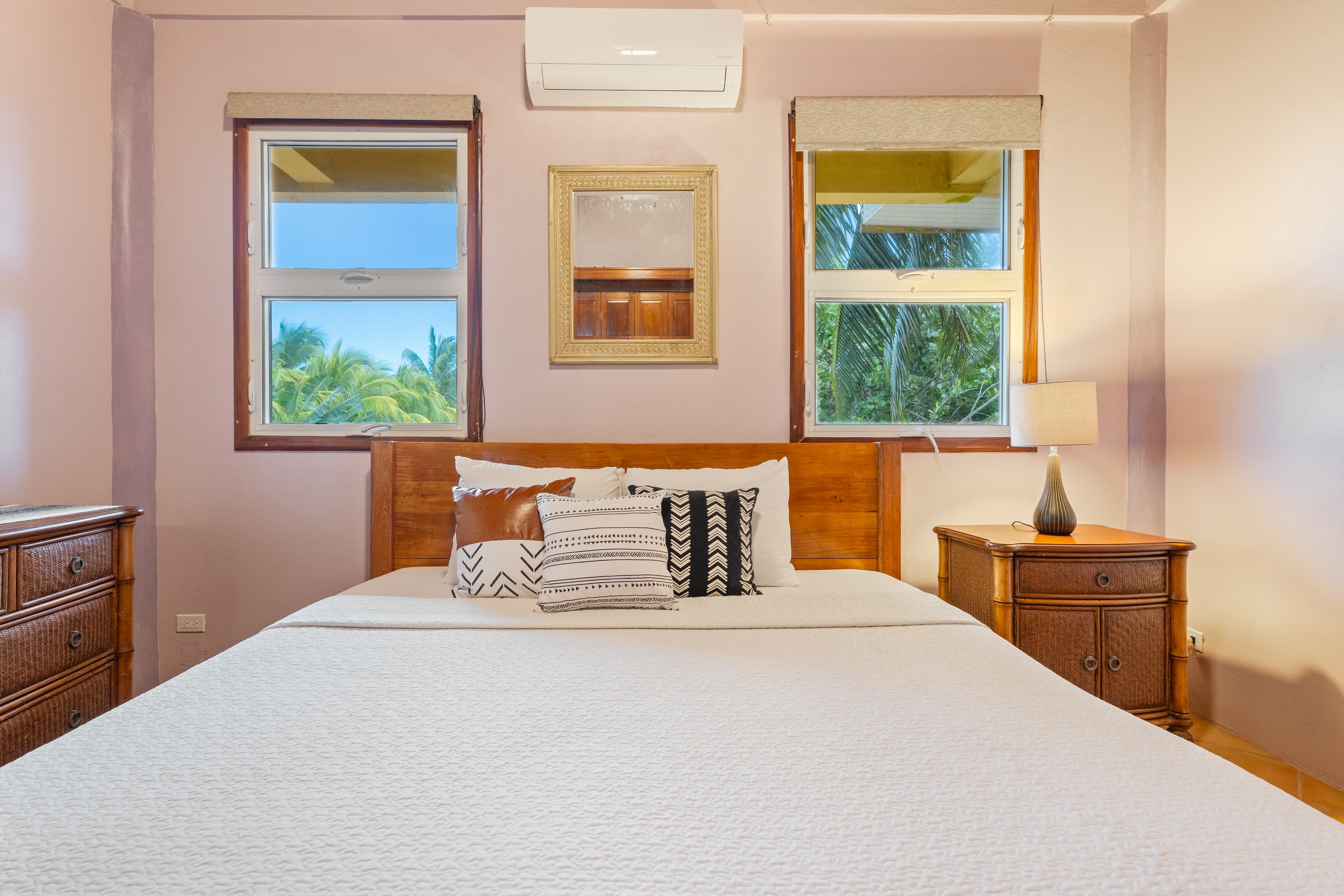 A bedroom with light lavender walls featuring a king bed with a wooden headboard and white textured linens adorned with several patterned accent pillows. The room has two windows offering views of lush greenery, a wooden nightstand with a lamp on the right, and a matching wooden dresser partially visible on the left.