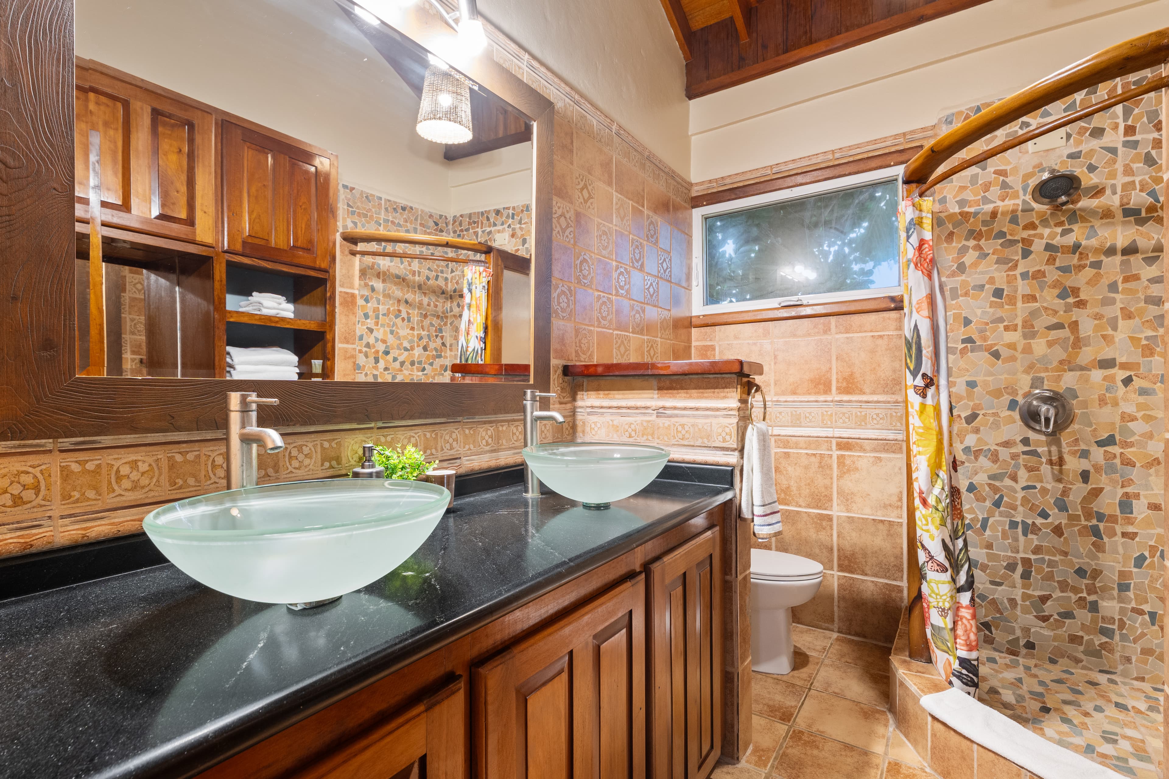 A spacious bathroom with warm-toned tiling and a high, dark wood ceiling. It features a double vanity with a dark countertop and two frosted glass vessel sinks positioned under a large wood-framed mirror. To the right, a walk-in shower is decorated with colorful mosaic tiles and a patterned shower curtain pulled to the side.