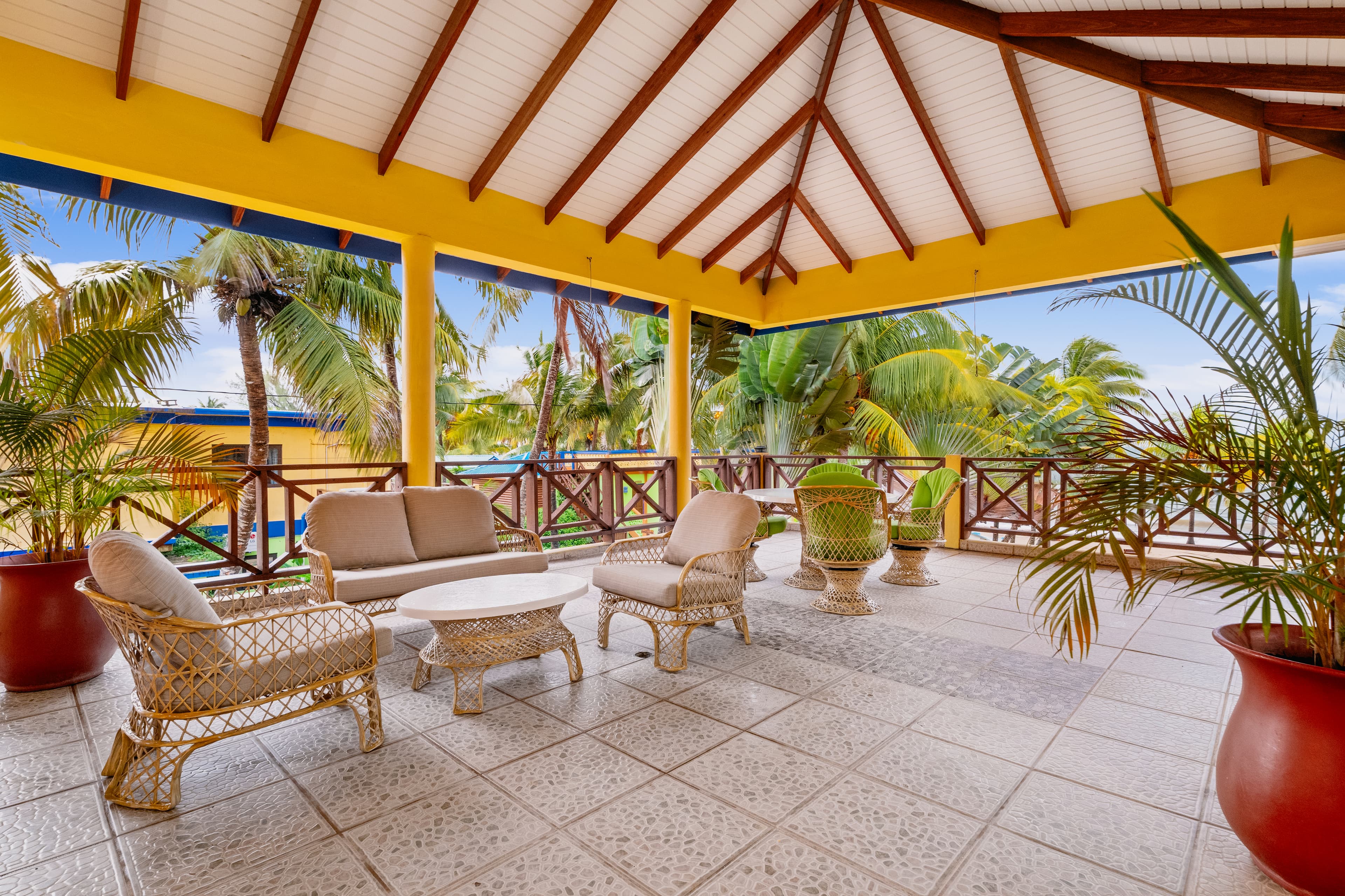 A spacious, covered outdoor patio with a vaulted white-beamed ceiling and tiled flooring. The area is furnished with a wicker lounge set featuring cream-colored cushions and a matching coffee table. In the background, a dining area with green chairs is visible, all overlooking a tropical landscape of palm trees under a bright blue sky.