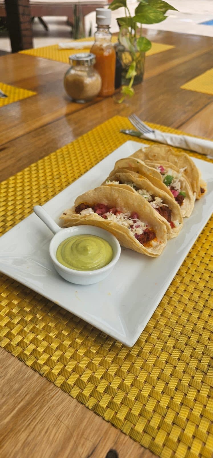 A plate of five tacos with various fillings and a small bowl of green sauce on a textured table mat.