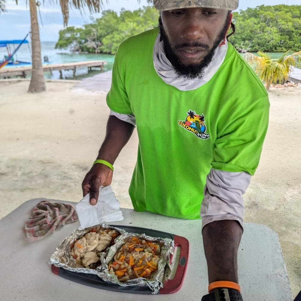 A person in a green shirt prepares food in aluminum foil on a table by the water.
