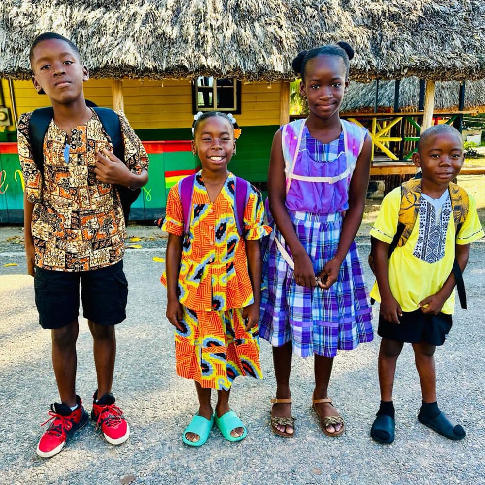 Four children in colorful outfits stand together in front of a straw-roofed building.