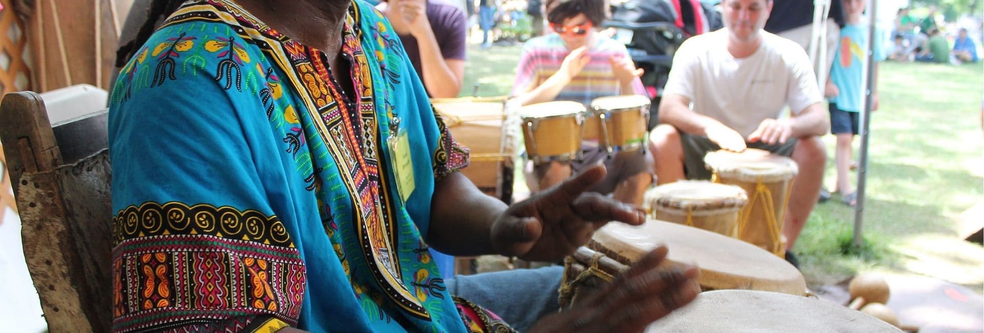 A musician plays drums in a colorful shirt while others join in the background.