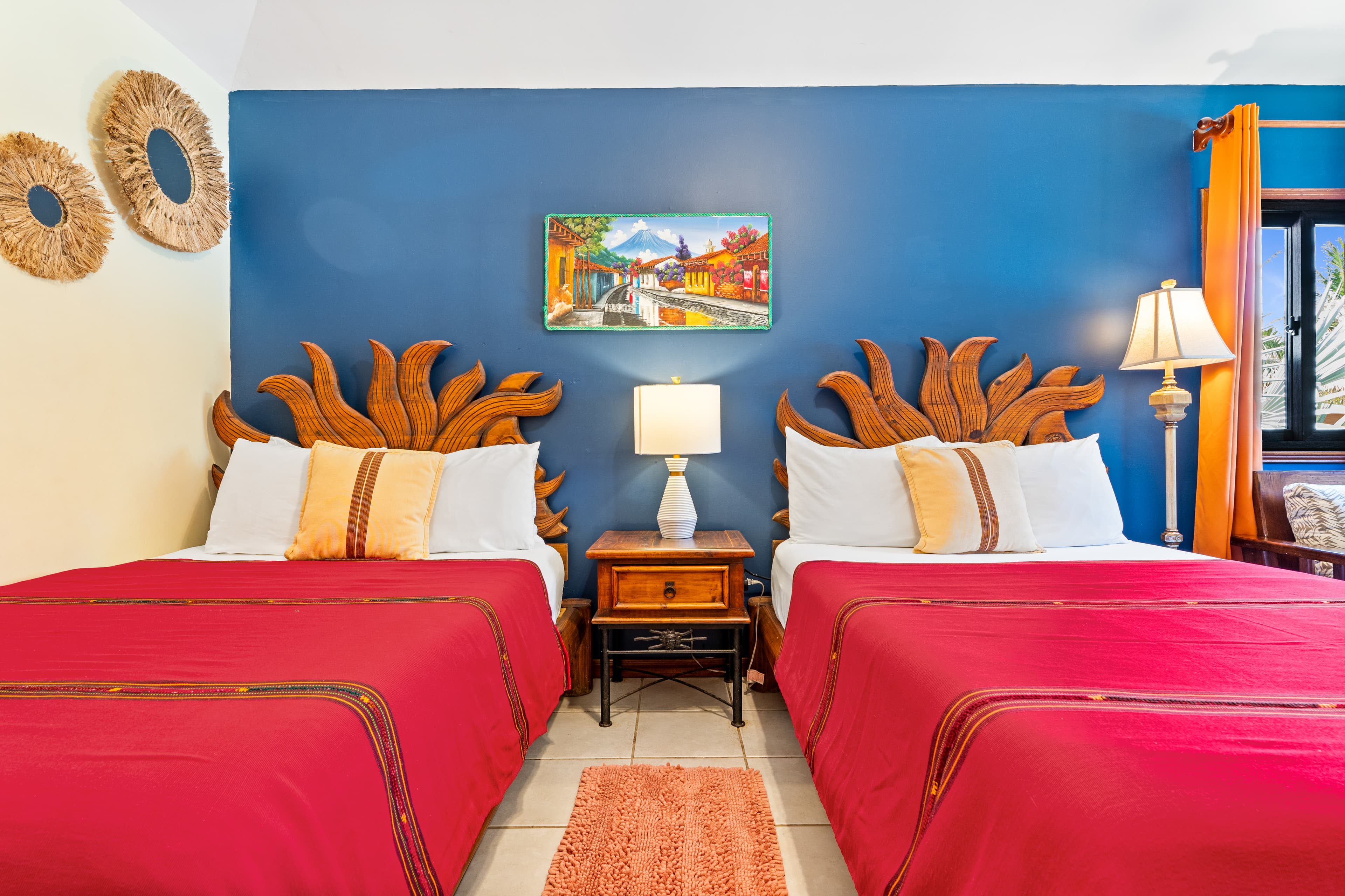 A vibrant bedroom featuring two full beds with bright red covers and unique, sunburst-shaped wooden headboards set against a deep blue accent wall. A colorful landscape painting hangs between the beds, and two woven circular wall decorations are mounted on the adjacent light-colored wall.